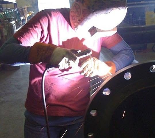 Person wearing gloves and a mask working with a welding tool on a circular metal object, illuminated by bright light.