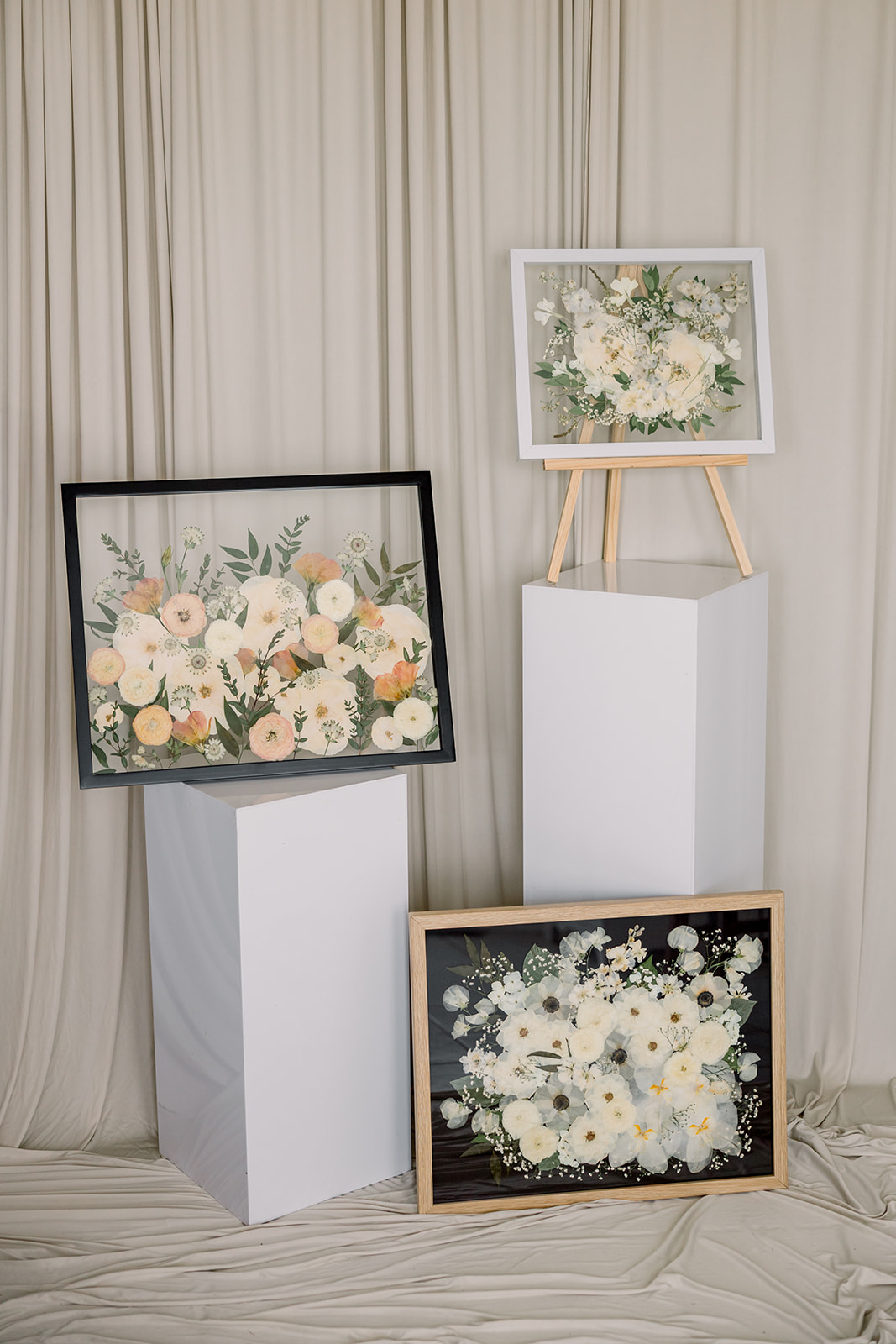 Three framed floral paintings displayed on white pedestals and a stand against a cream curtain background.