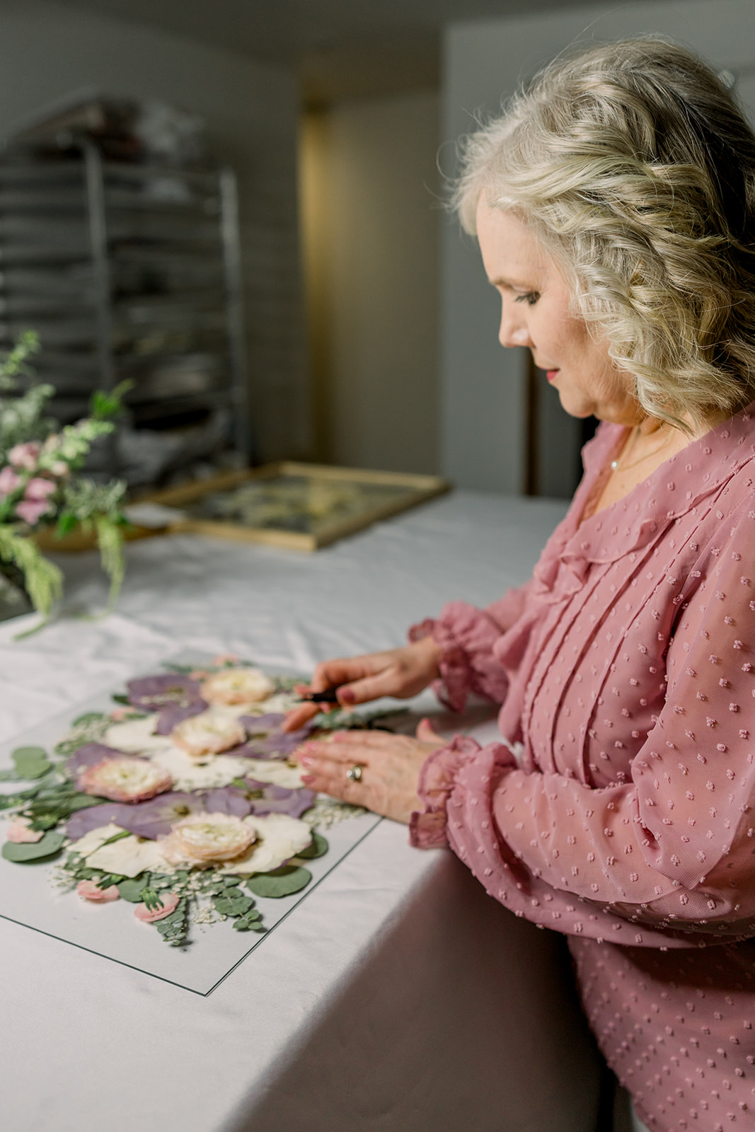 Older woman with curly gray hair arranging floral cutouts on a table, wearing a pink blouse, in a room with shelves.
