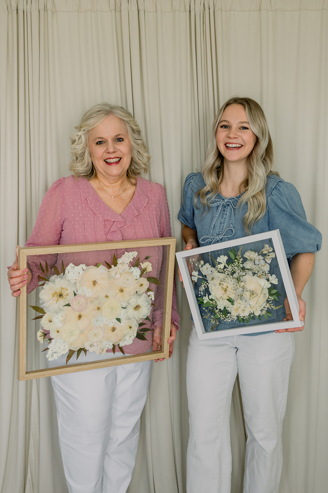 Two women holding framed floral arrangements, smiling, standing in front of a light-colored curtain.