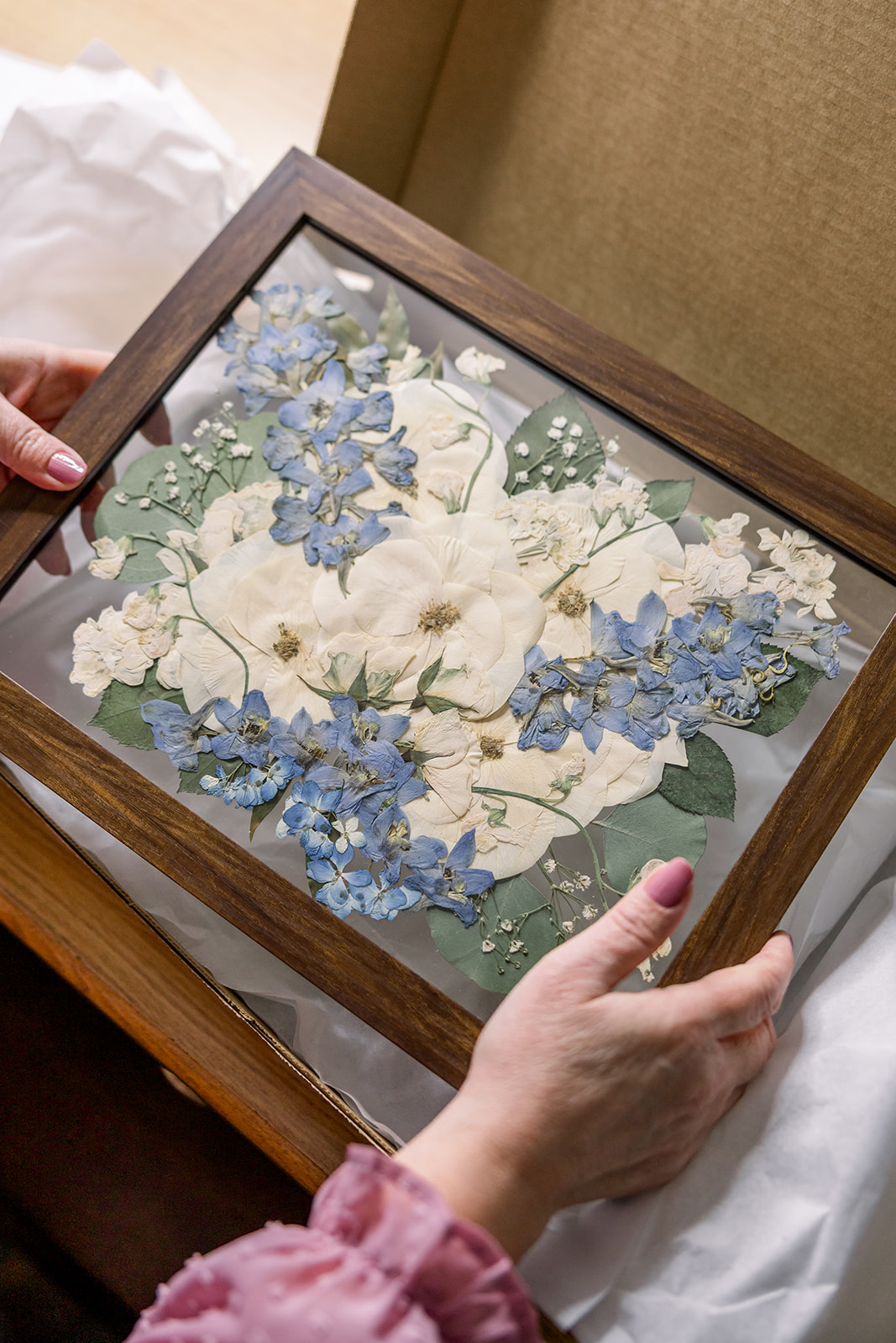 Person holding a framed floral artwork with white and blue flowers, wooden frame, on a bed with white bedding.