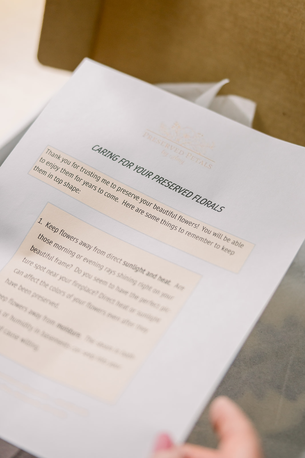 A document titled 'Caring for Your Preserved Floral' with blurred text, held by a person's hand, on a table.