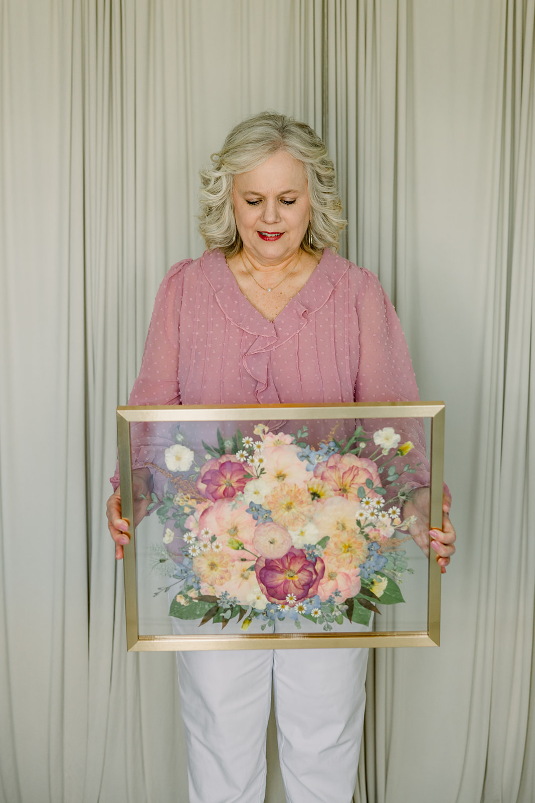 Older woman with gray hair in a pink sweater holding a floral painting, standing in front of light-colored curtains.