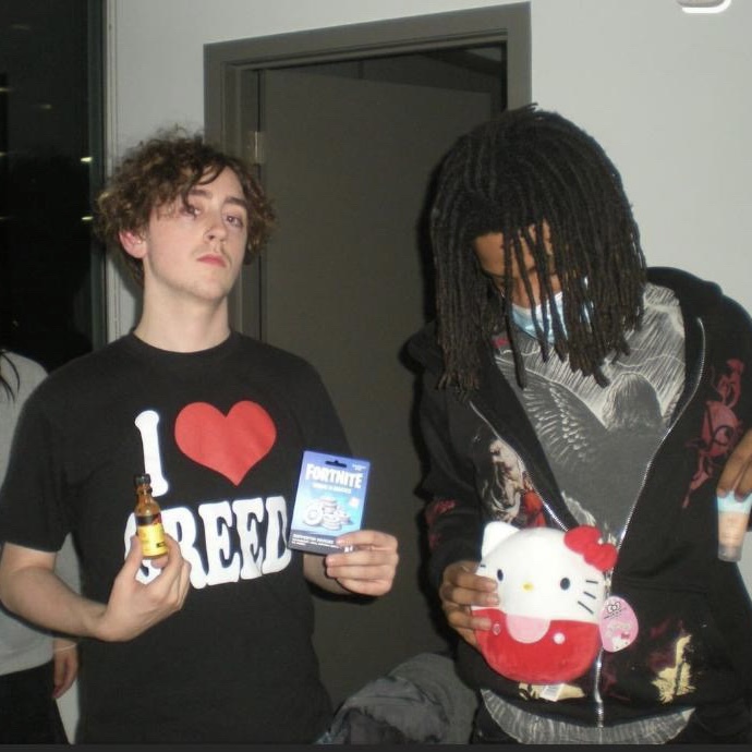 Two young men standing indoors, one holding a small bottle and a Fortnite card, the other holding a Hello Kitty plush toy.