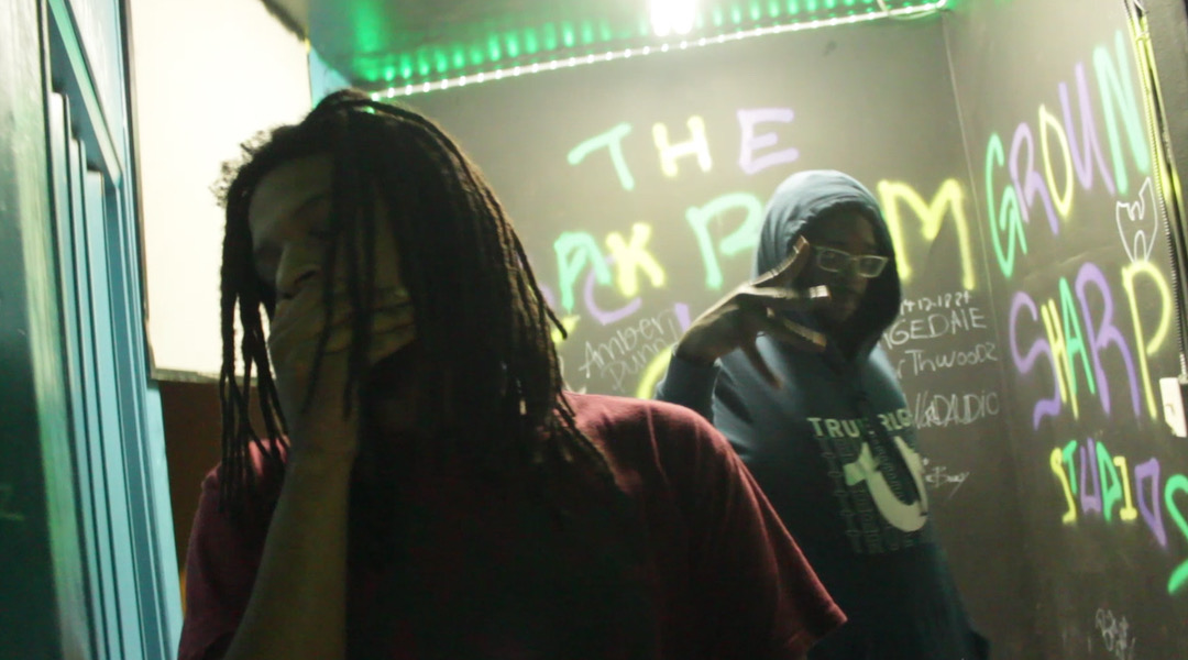 Two people in a dimly lit room with colorful graffiti on the wall behind them, one with dreadlocks and the other wearing glasses and a hoodie.