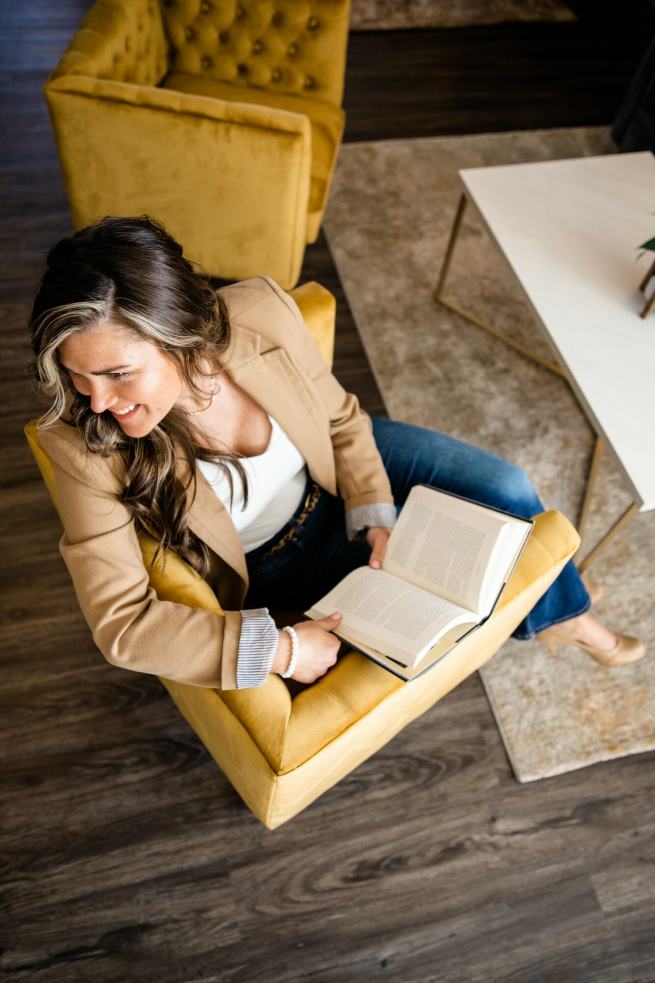 Woman sitting in a yellow armchair reading a book, with a white table nearby, in a cozy room.