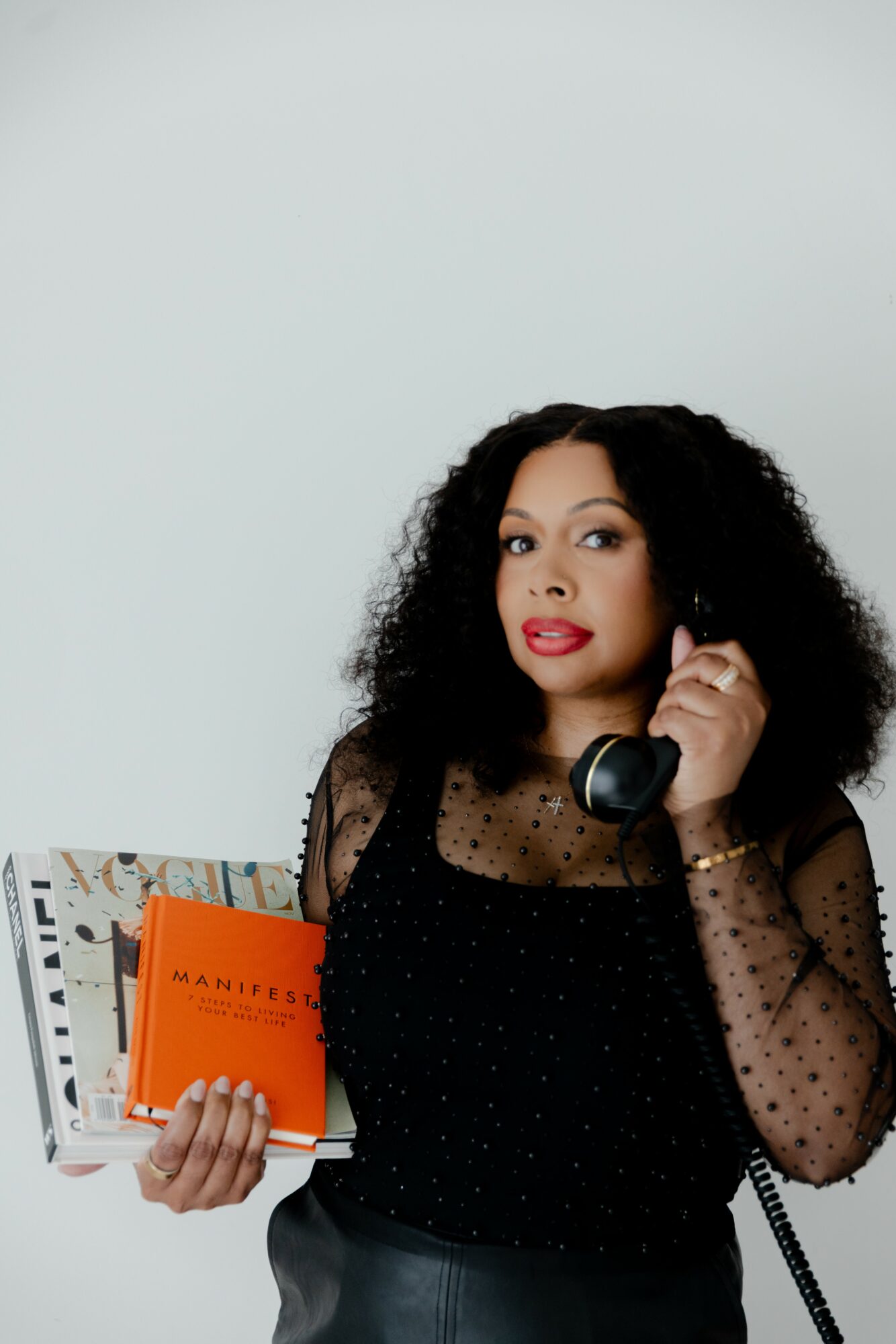 Woman with curly hair holding a phone to her ear and a stack of books, wearing a black sheer top, against a plain background.