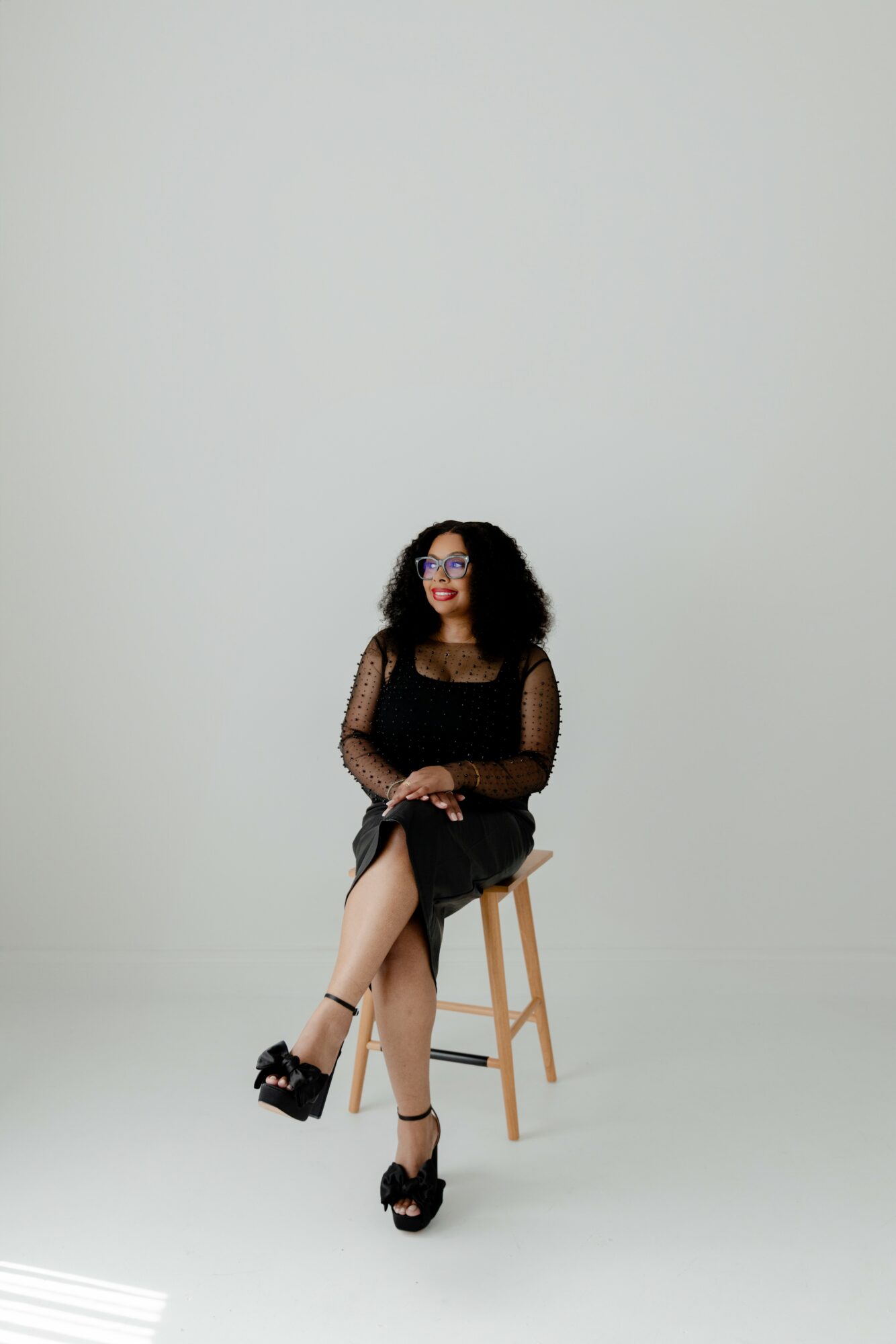Woman with curly hair sitting on a stool, wearing a black dress and glasses, against a plain light background.