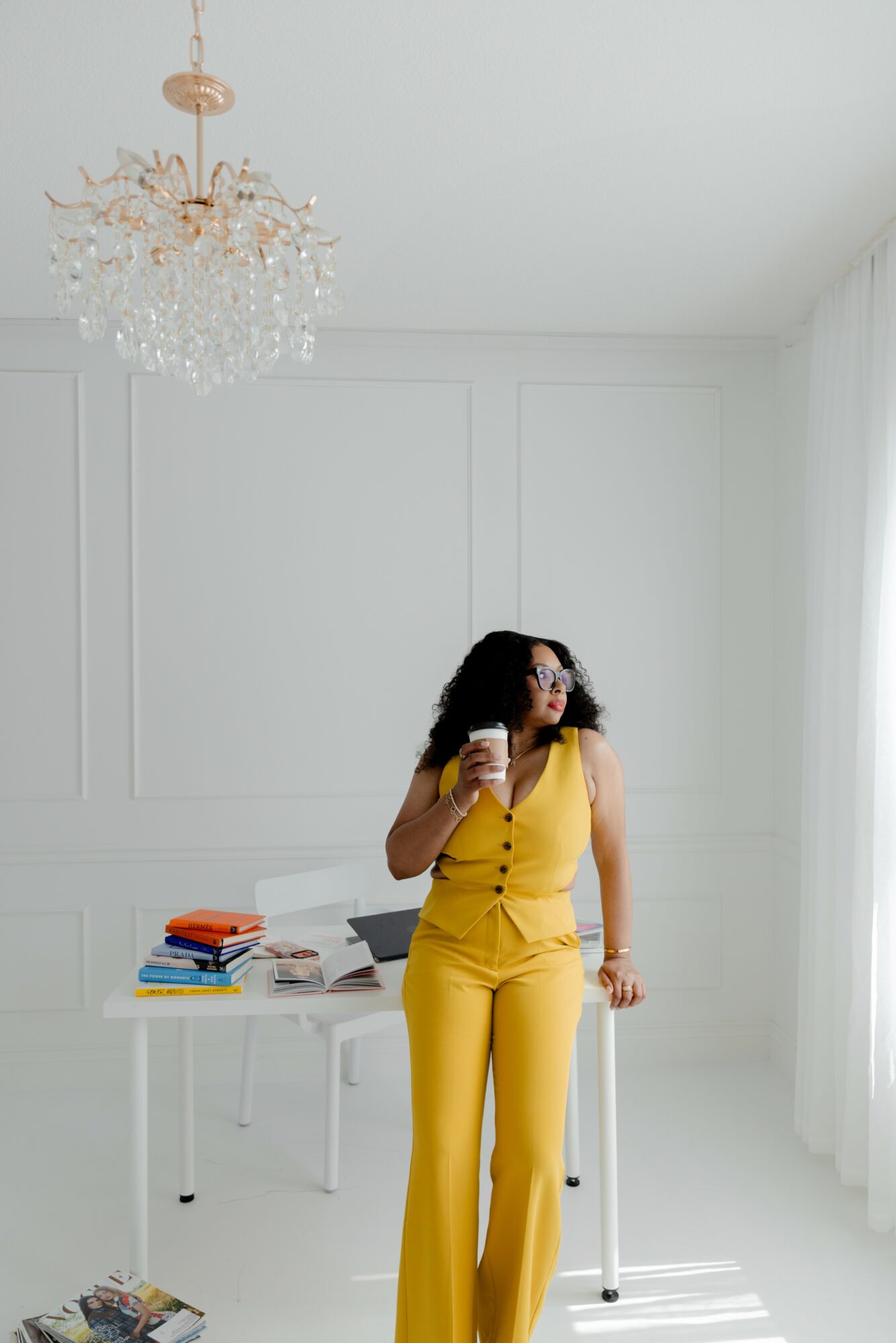Woman in yellow outfit holding a glass, standing near a table with books and papers, in a bright room with white walls and curtains.