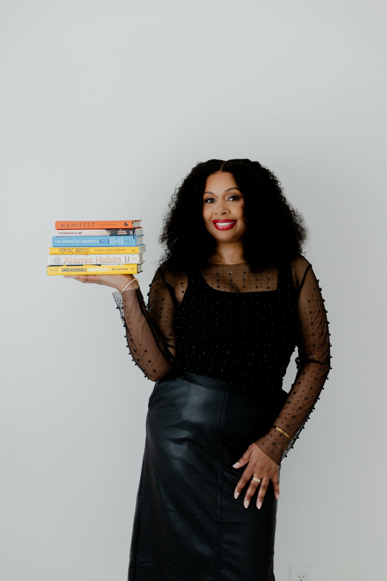 Woman with curly hair holding a stack of books, smiling, wearing a black sheer top and skirt, standing against a plain background.