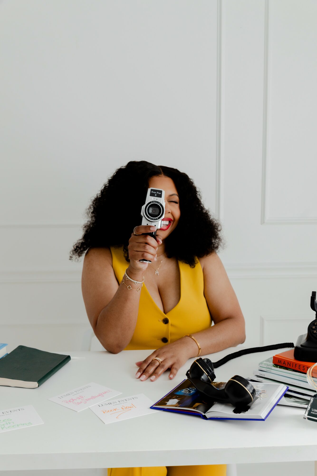 Woman with curly hair in yellow top holding a camera, sitting at a white desk with papers and office supplies.