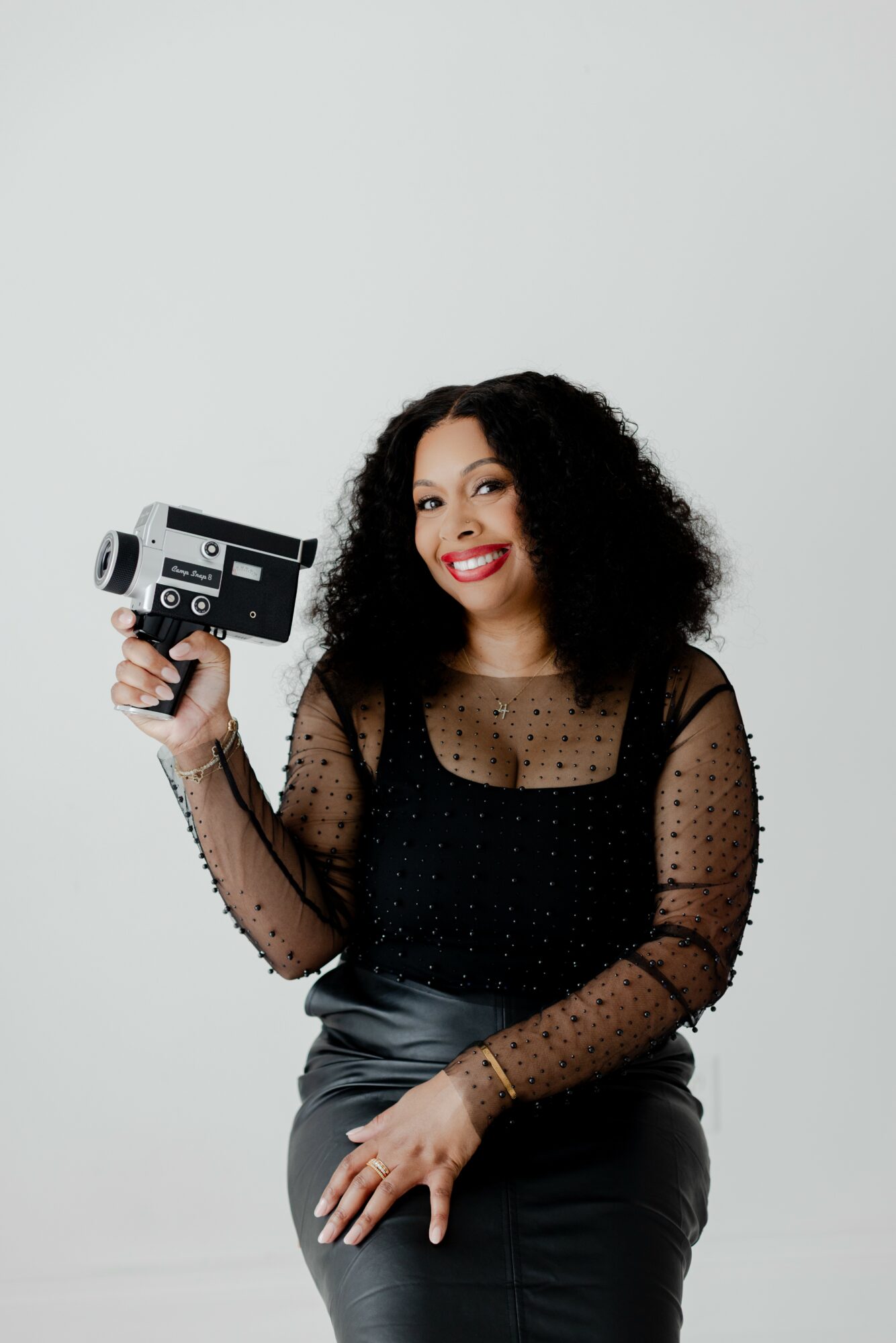 Woman with curly hair holding a vintage film camera, smiling, wearing a black sheer polka dot top and leather skirt.