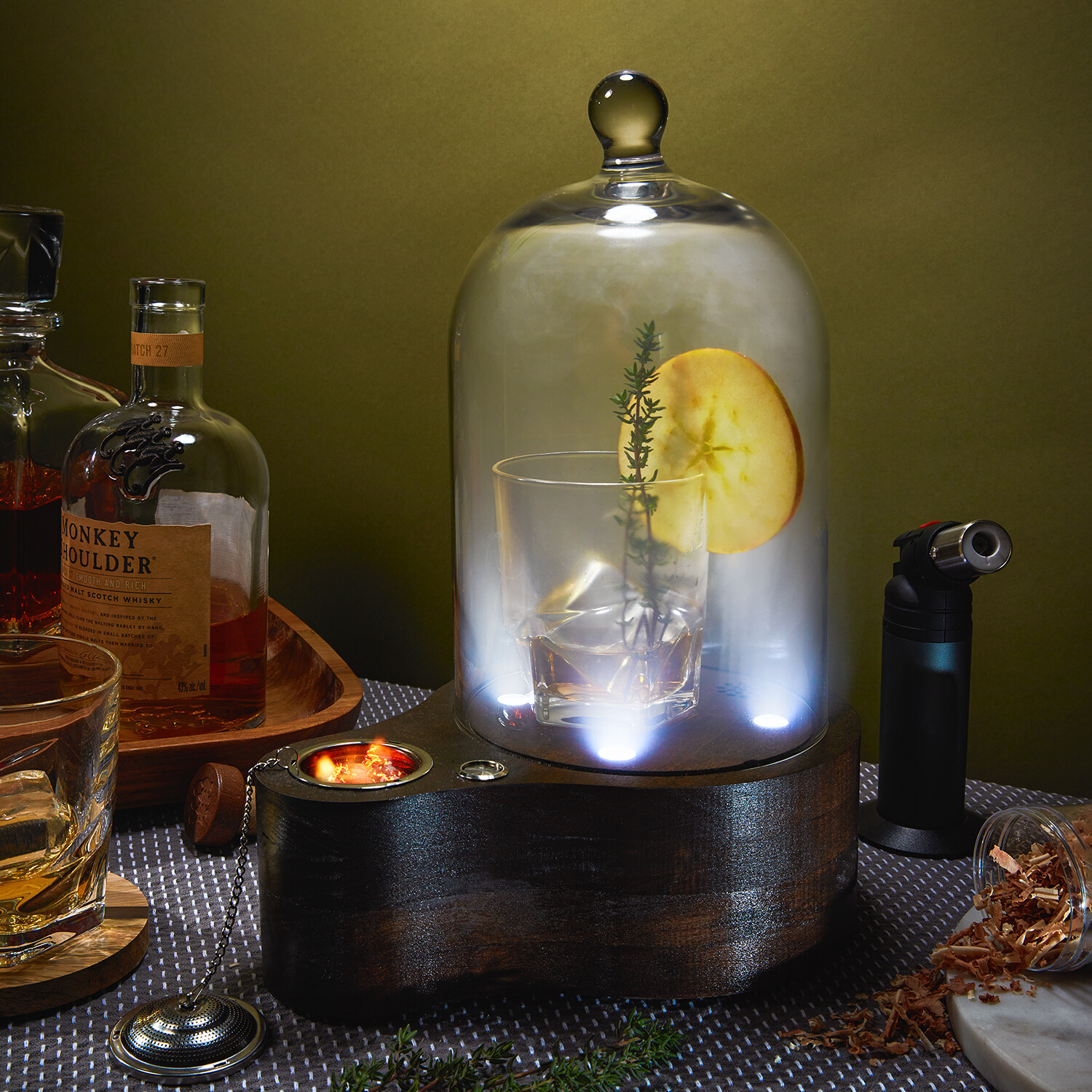 Glass container with lemon slice and herbs, water inside, on a dark wooden base, surrounded by bottles and objects.