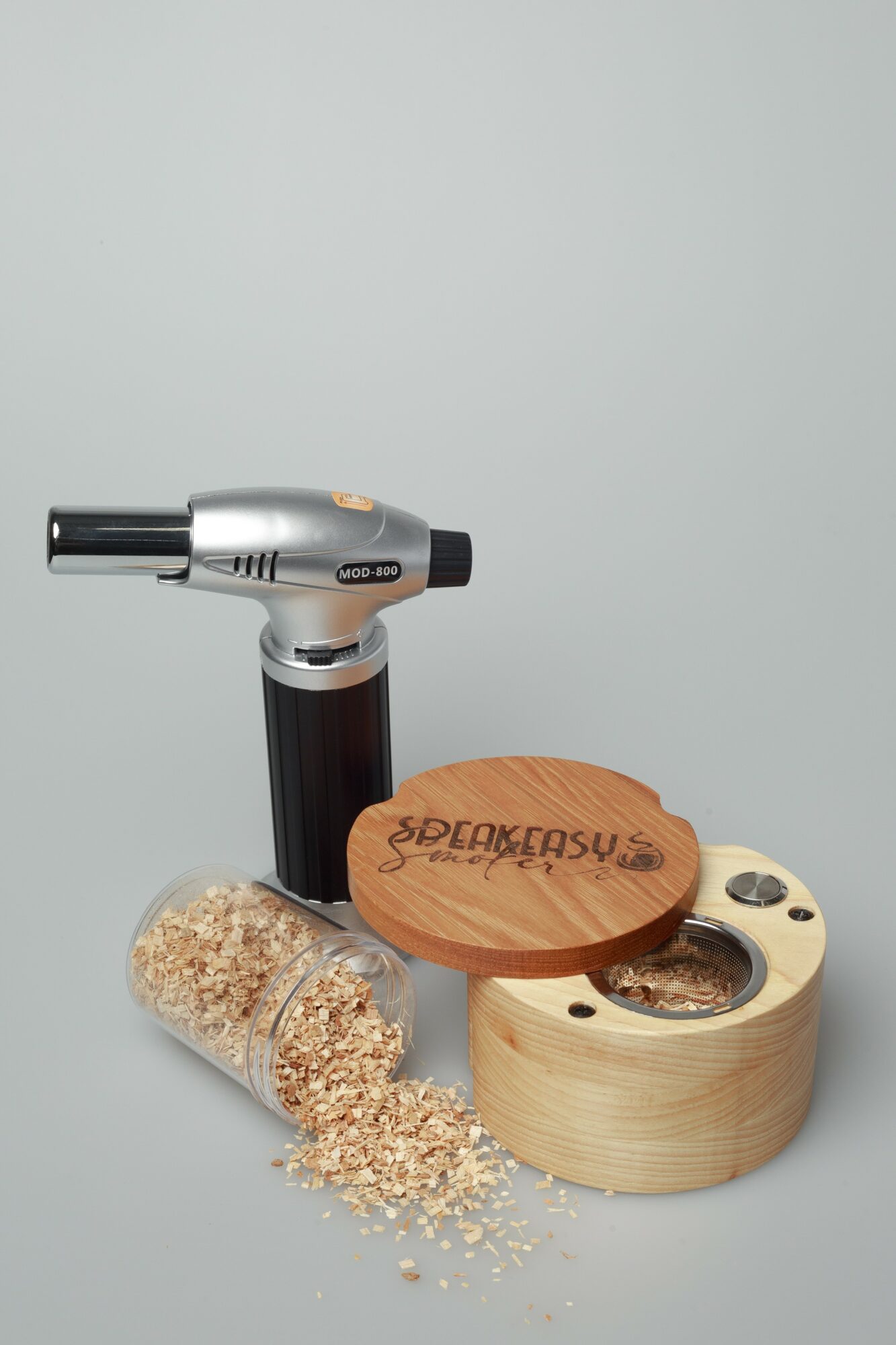 A wooden container with a lid, a small wooden disc, a black and silver tool, and a glass jar with oats on a light background.