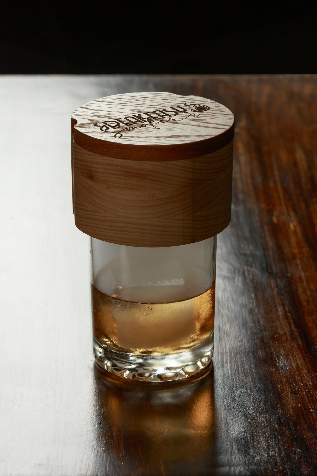 Glass with liquid and a wooden lid on a dark wooden surface, background is dark and blurred.