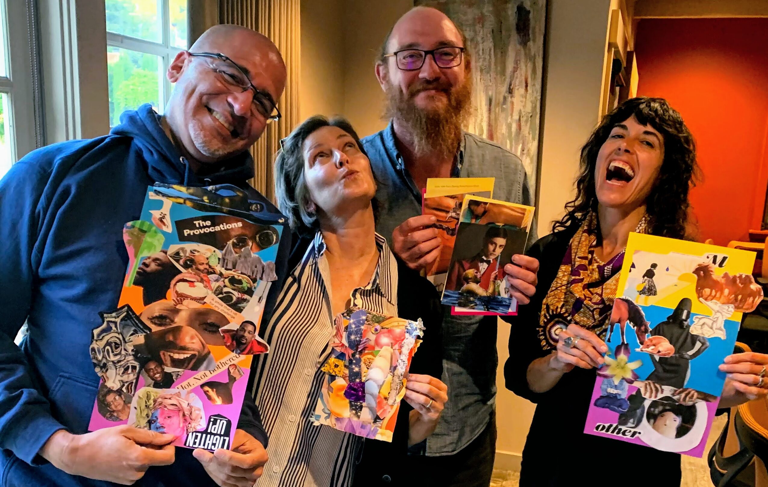 Four people smiling and holding colorful comic books indoors, with a window and curtains in the background.