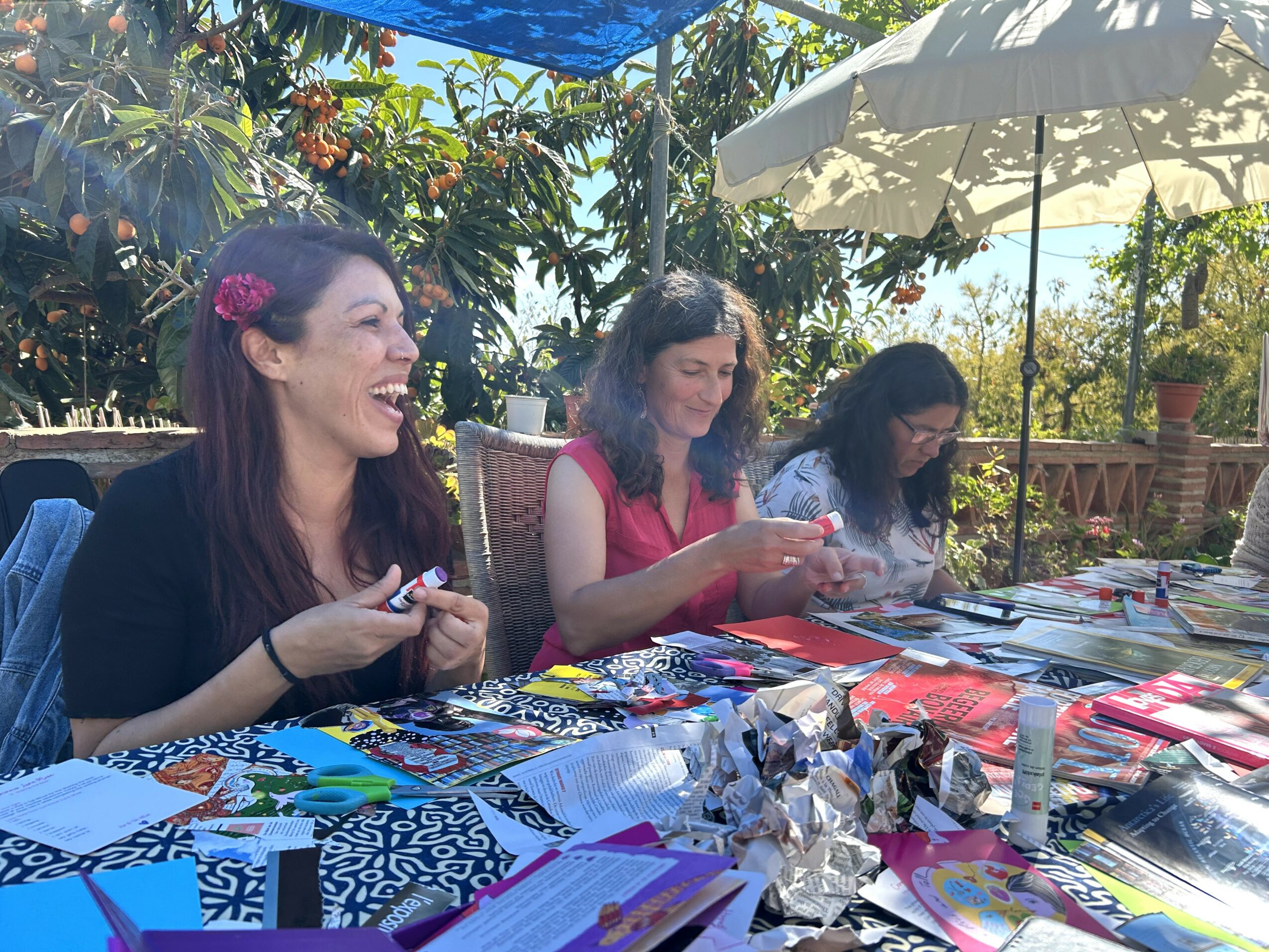 Three women sitting at a table outdoors, smiling and looking at their phones or books, with colorful items on the table.