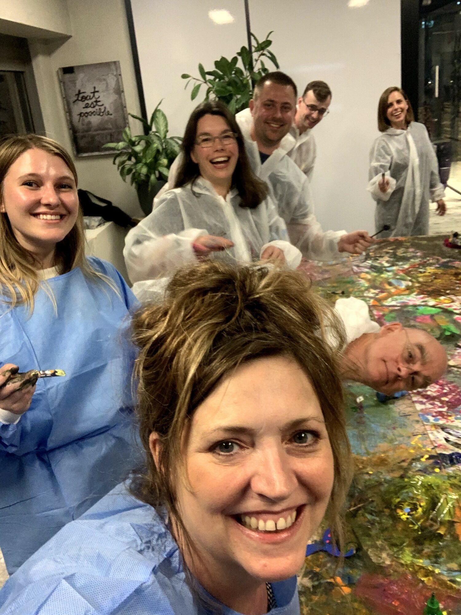 Group of people smiling and posing around a colorful table, some wearing protective smocks, in an indoor setting.