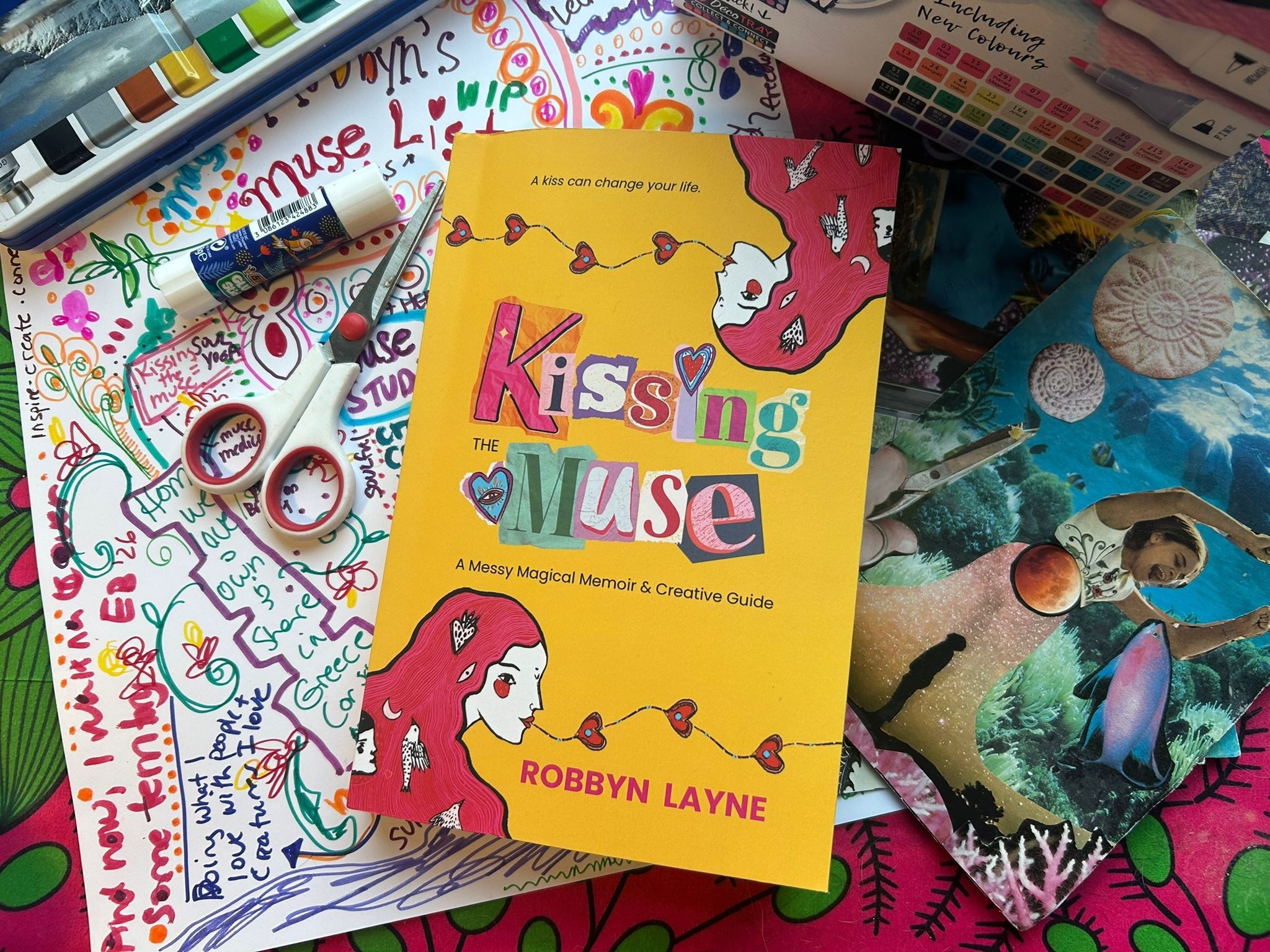 Colorful book titled 'Kissing the Muse' by Robbyn Layne, surrounded by art supplies, notebooks, and a photo of a dog with sunglasses.