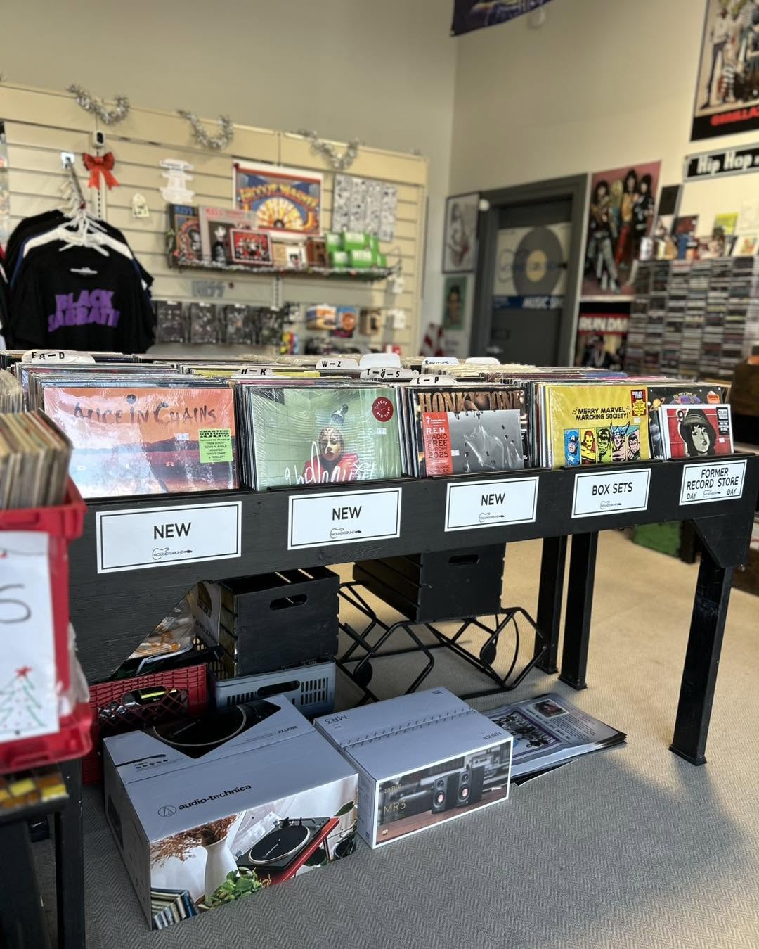 Display table with vinyl records and boxes in a music store, shelves with CDs and posters in the background.