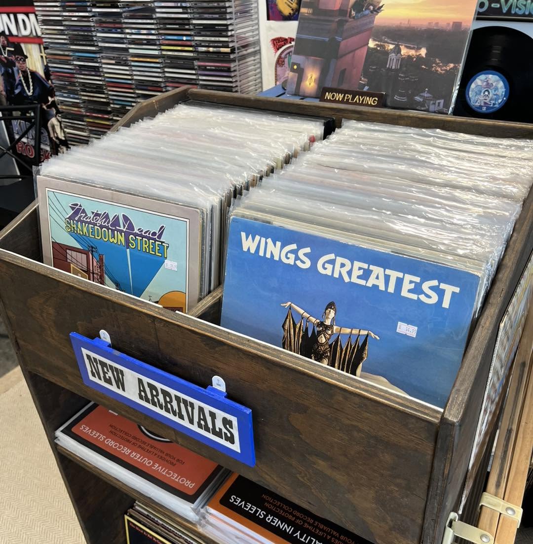Wooden display box with vinyl records, including 'Wings Greatest' and 'New Arrivals' sign, in a store.
