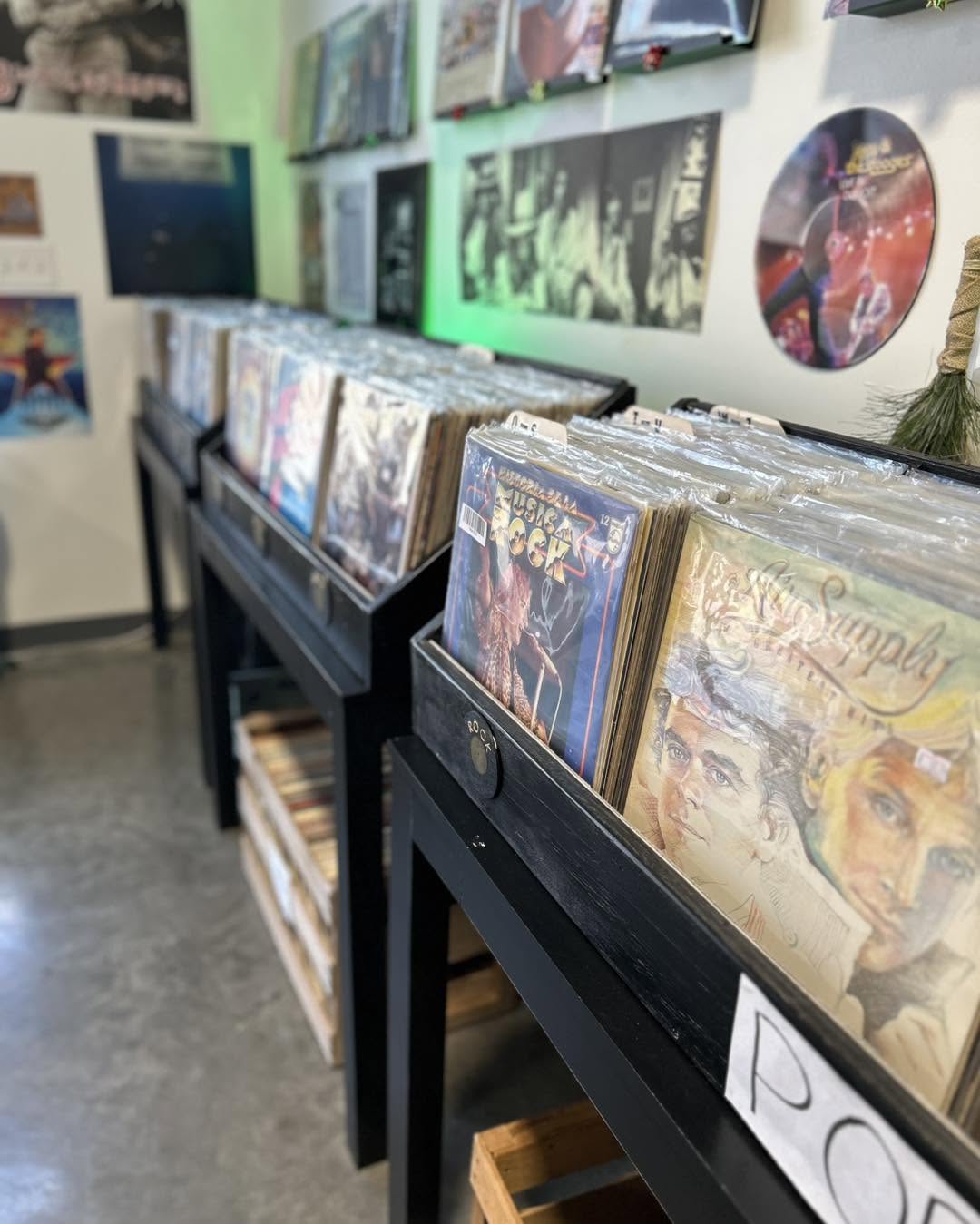Records displayed on black shelves with album covers visible, wall decorated with posters and artwork, floor visible in the background.