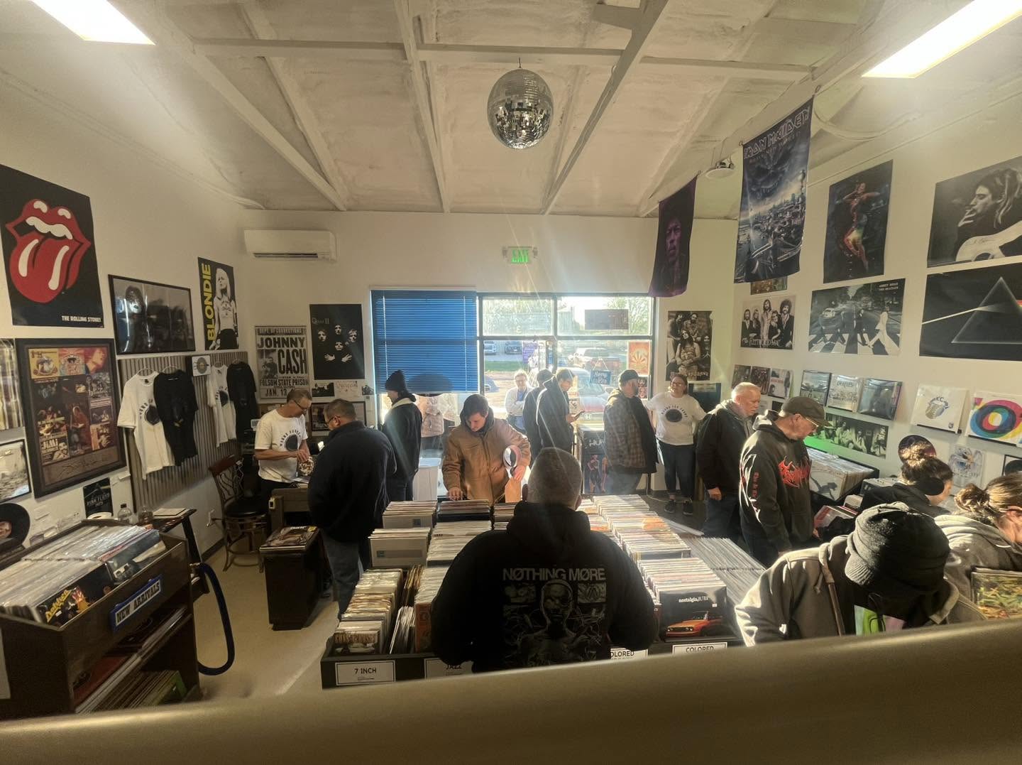 People browsing vinyl records in a store with posters and artwork on the walls, ceiling, and window in the background.