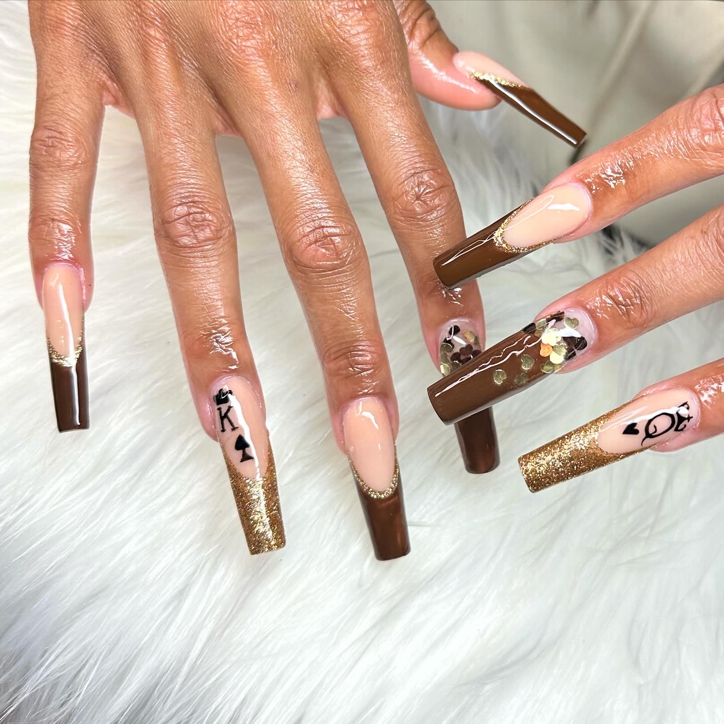 Close-up of hands with decorated long nails, some with gold, brown, and black designs, on a white furry surface.