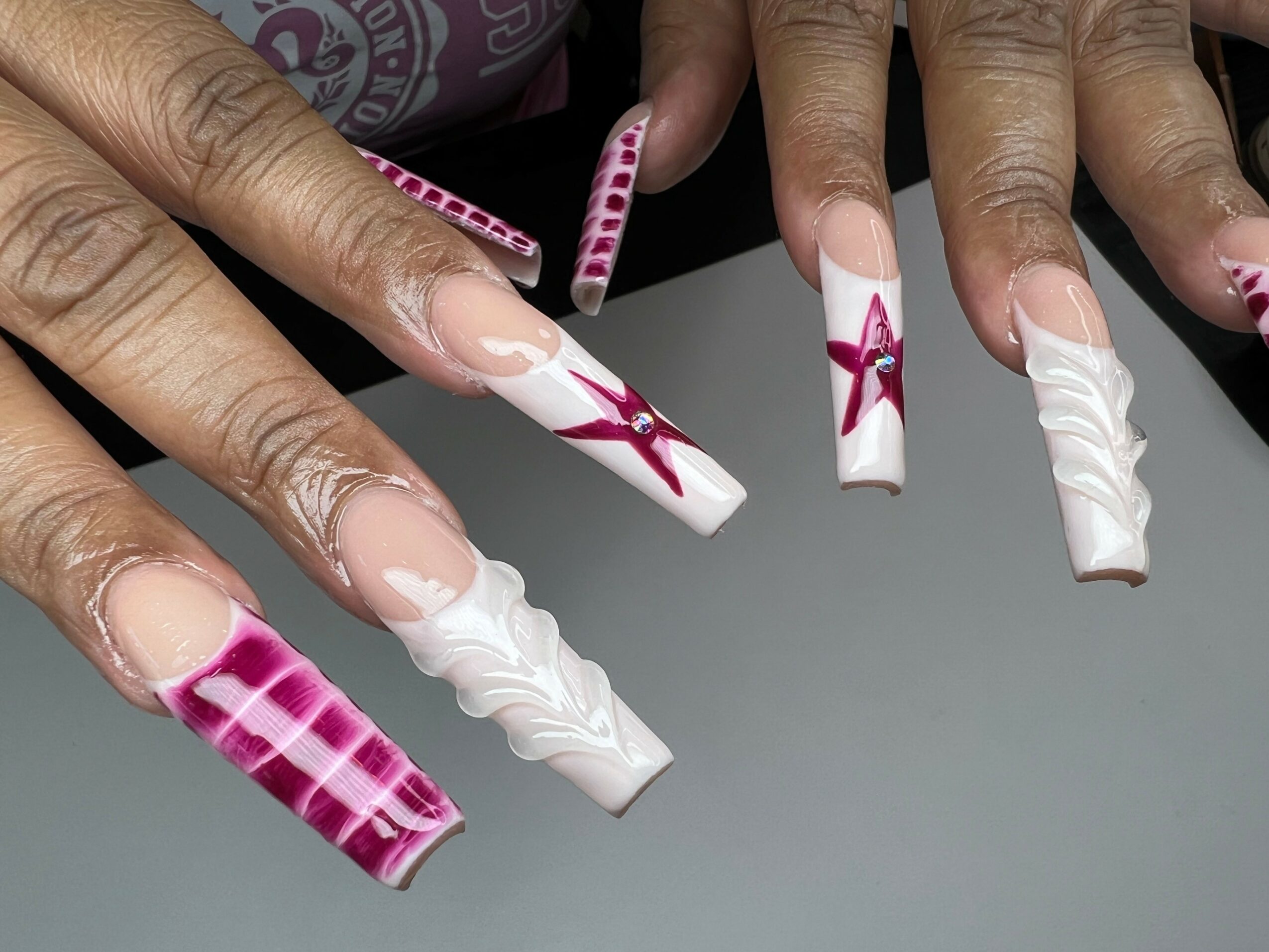 Hands with decorated long nails, featuring pink and white patterns and bows, resting on a gray surface.