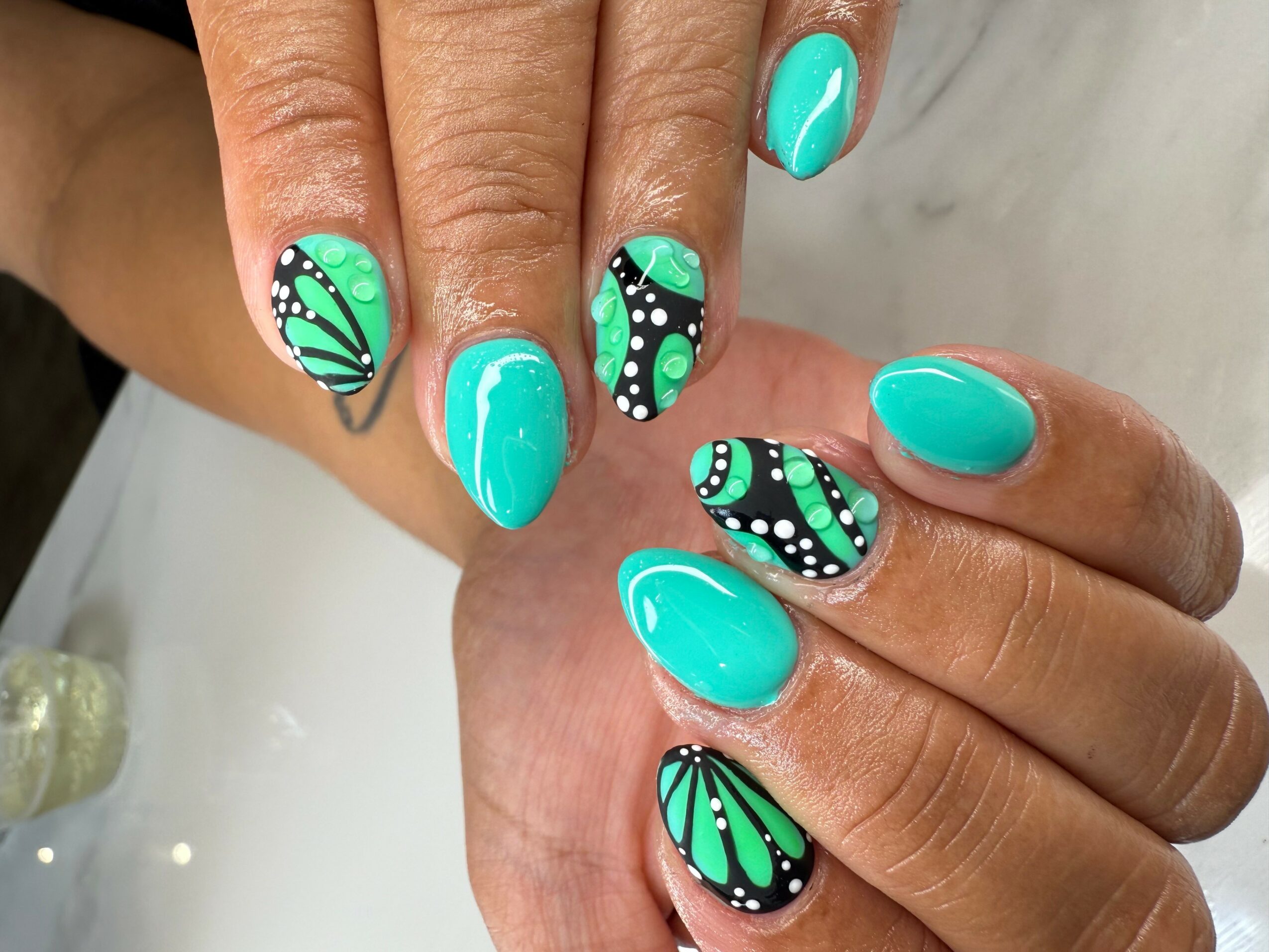 Hand with turquoise and black patterned nail polish, featuring dots and lines, on fingernails and thumb.