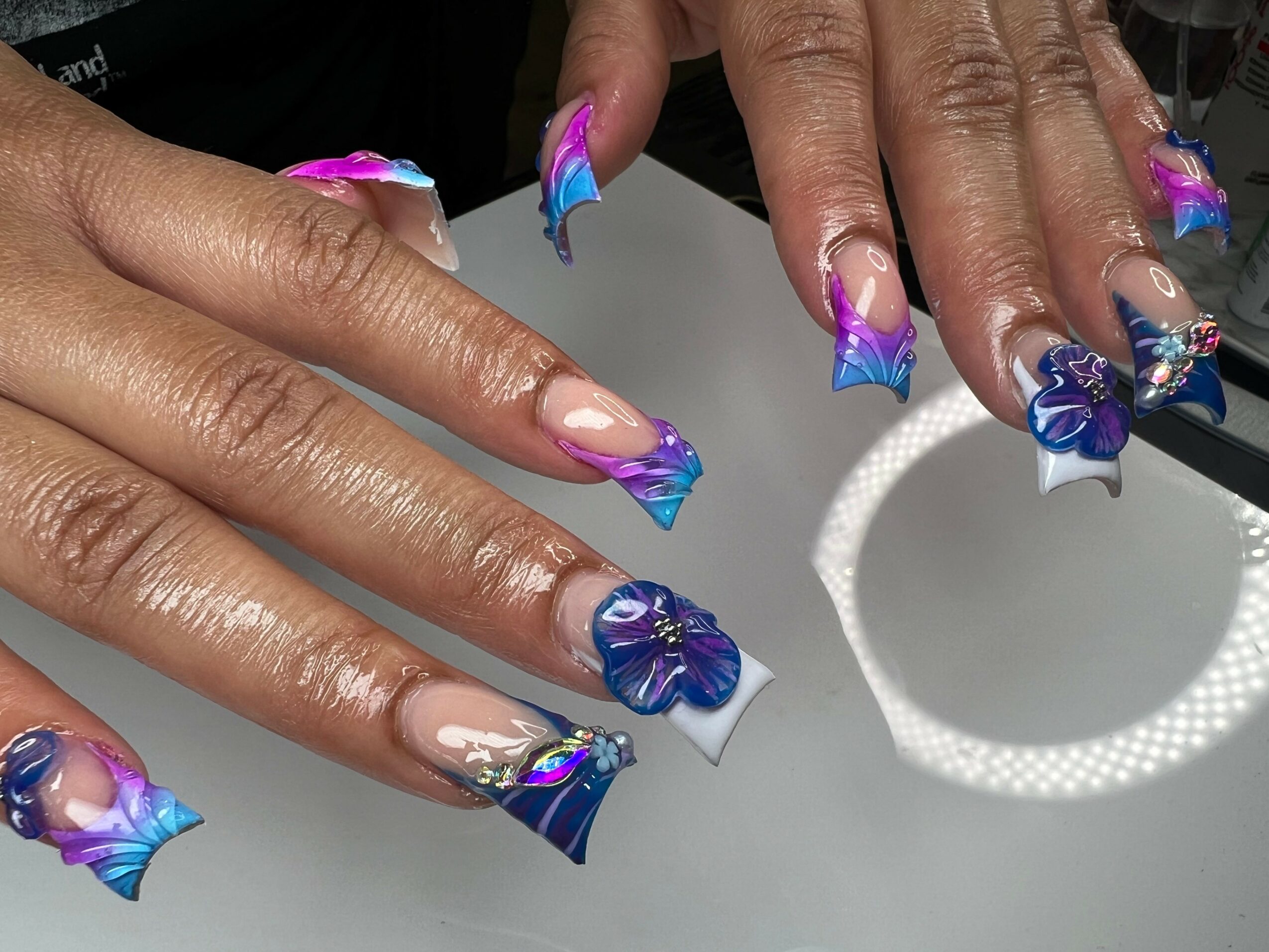Hands with decorated nails featuring blue, pink, and purple designs, resting on a white surface.