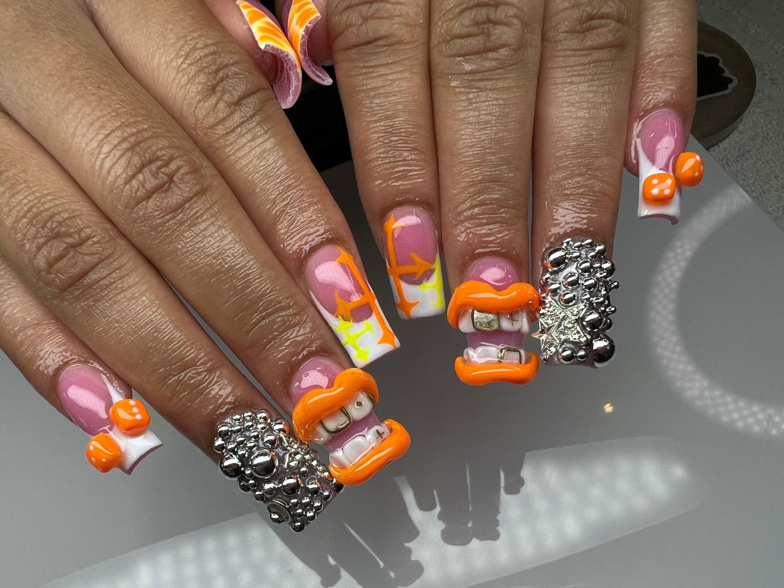 Hands with decorated nails featuring orange, pink, and purple colors, some with rhinestones and 3D designs, and rings with rhinestones.