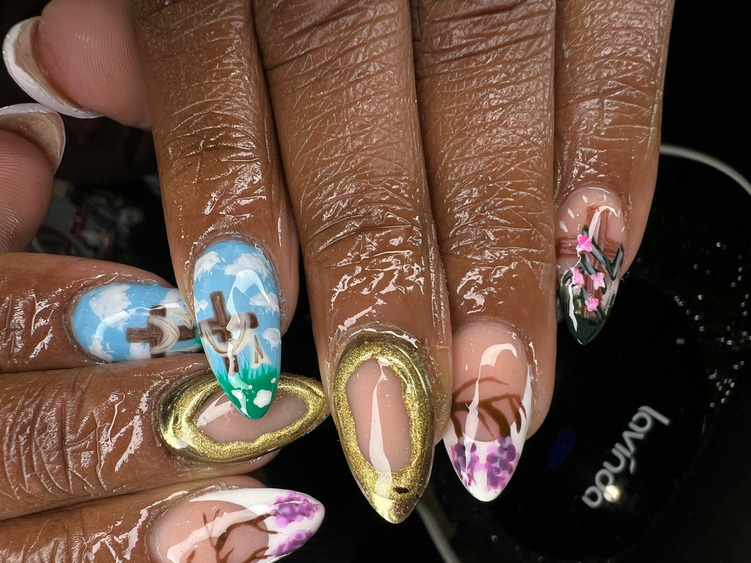 Close-up of hands with colorful, detailed nail art on fingernails, including floral and abstract designs, with gold accents.