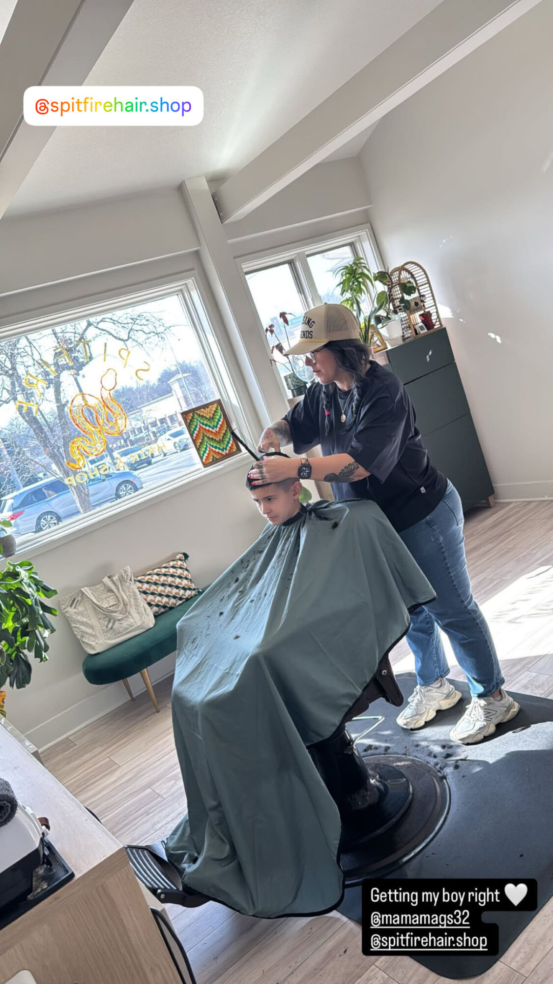 Person receiving a haircut in a salon, wearing a hat and mask, with a large window and plants in the background.