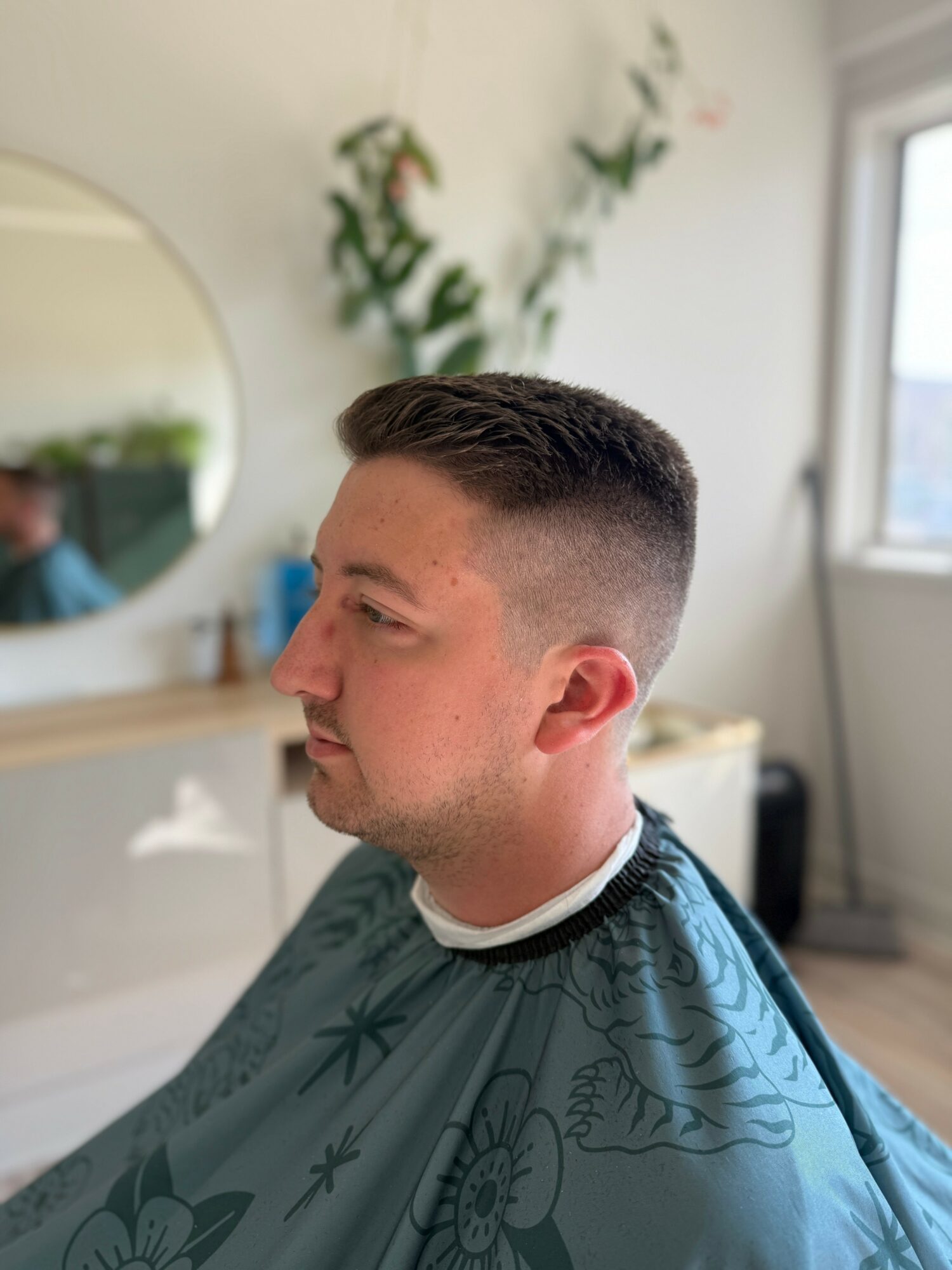 Young man with a fresh haircut sitting in a salon, wearing a cape, with a mirror and plants in the background.