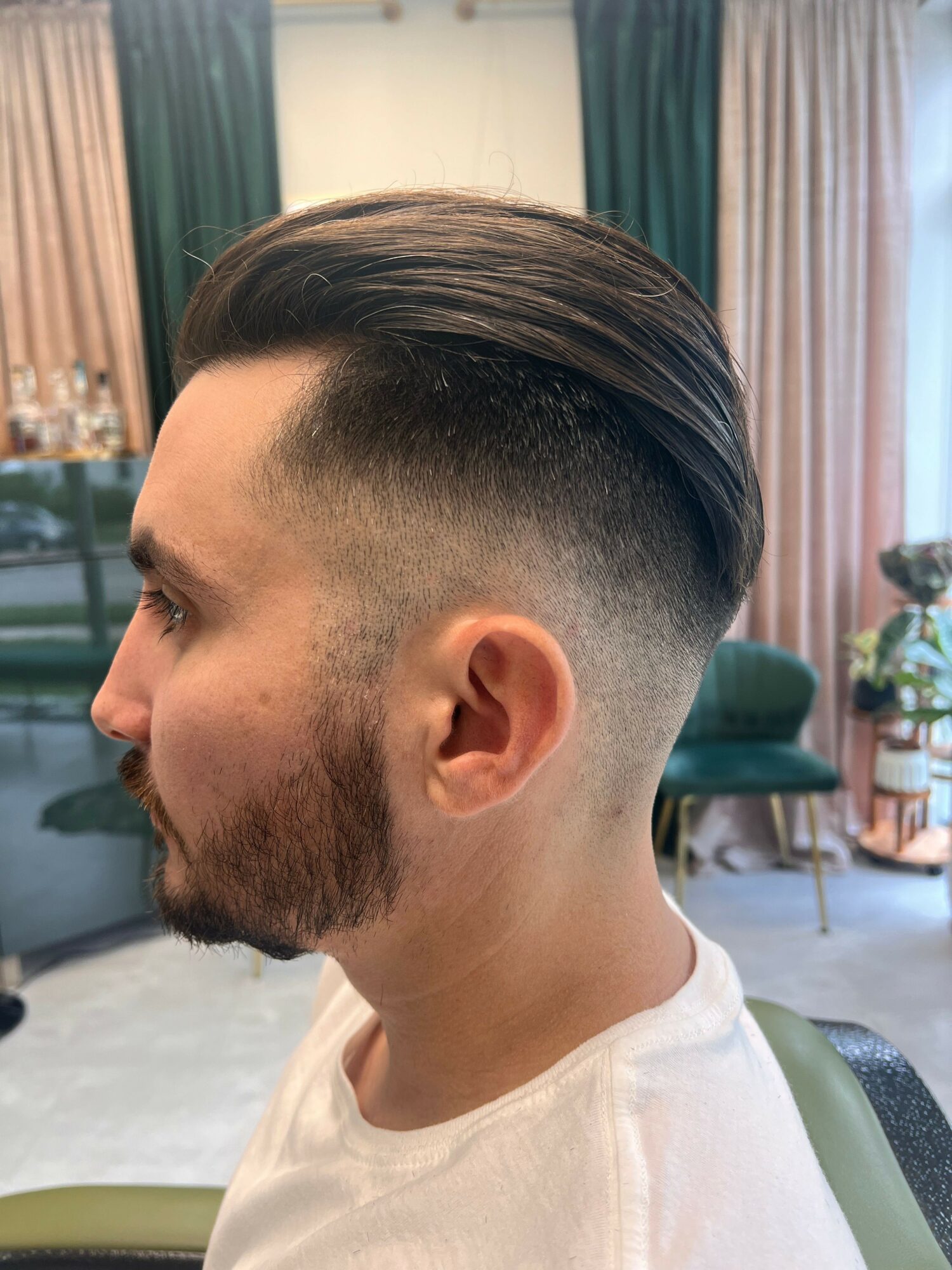 Side profile of a man with a styled haircut and beard, indoors with curtains and furniture in the background.