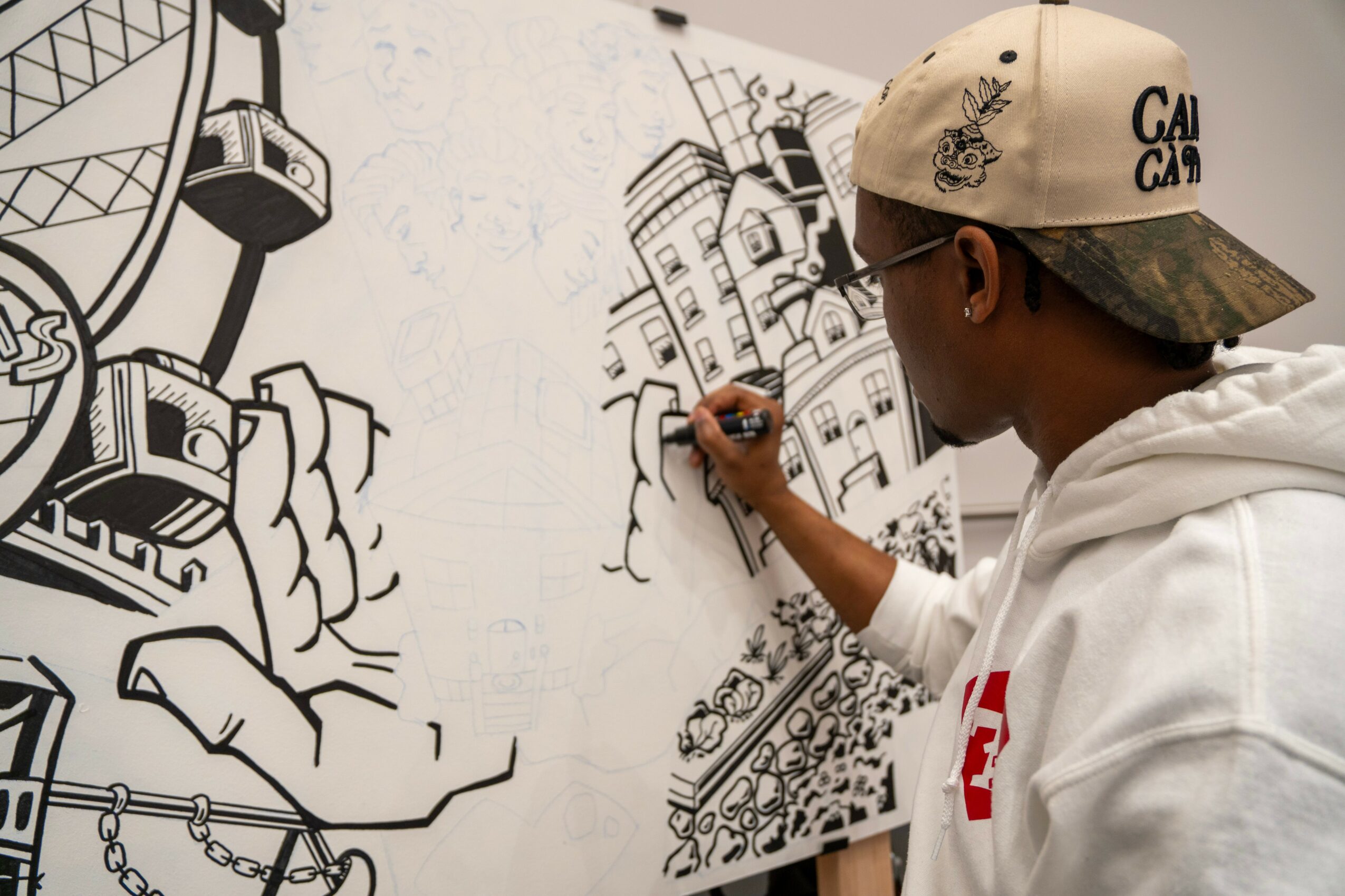 Person drawing on a wall with black marker, wearing a cap and glasses, with cityscape sketches in the background.