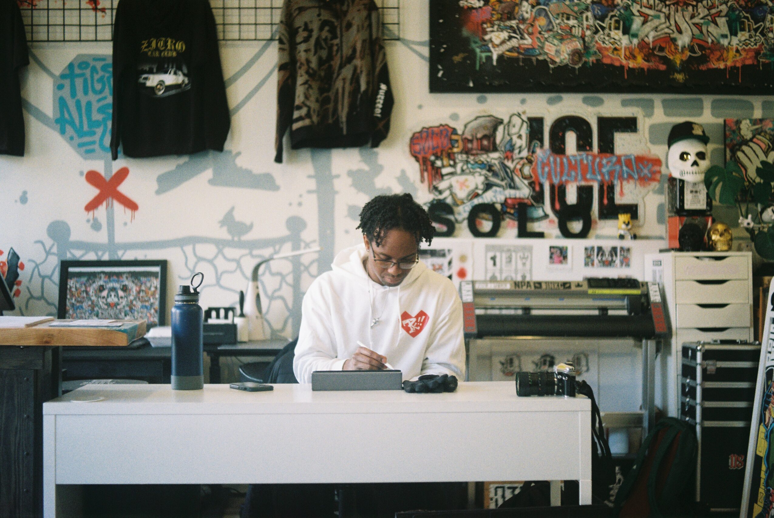 Person with glasses and curly hair sitting at a desk in a room with graffiti and artwork on the wall, writing or drawing.