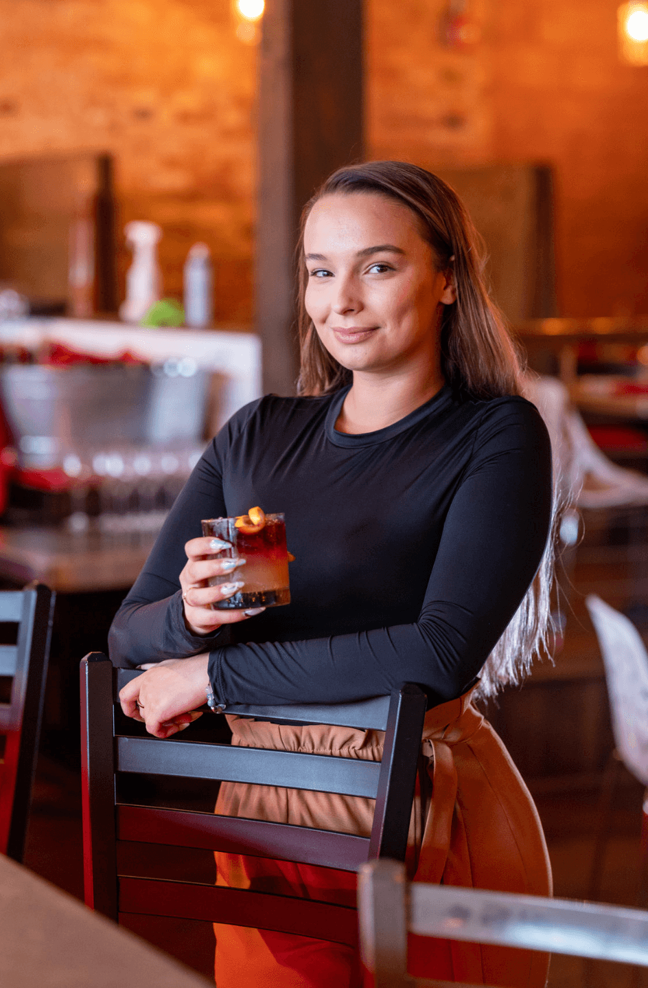 Young woman with long hair holding a drink, smiling in a restaurant or bar setting, brick wall background.