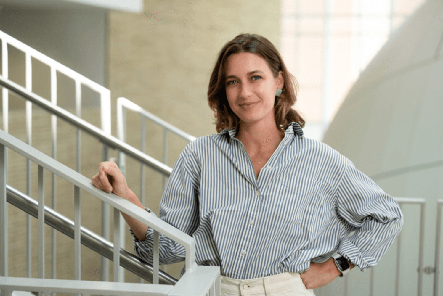 Woman with shoulder-length hair in a striped shirt standing on stairs outdoors, smiling, with modern architecture in background.