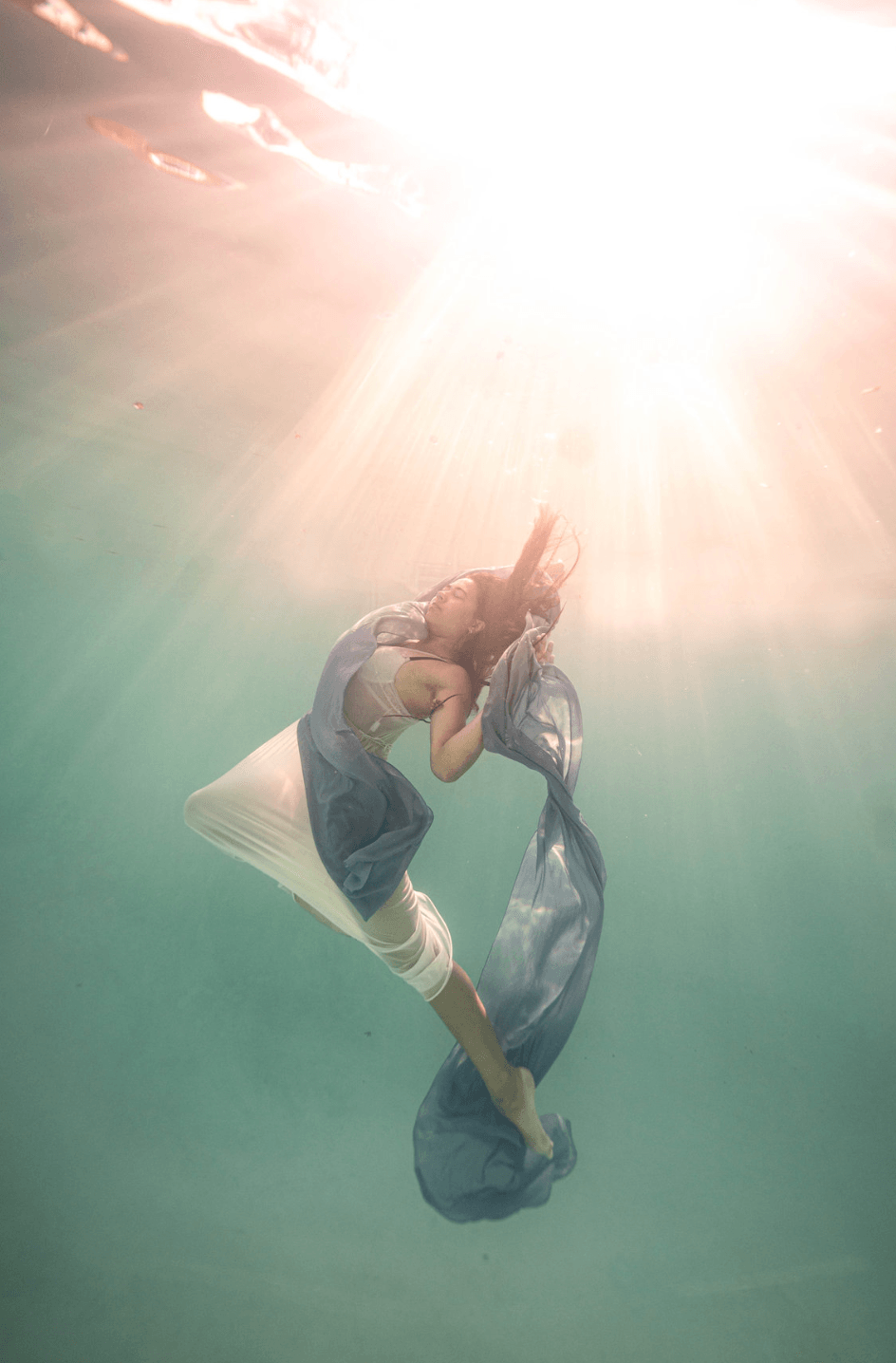Person underwater holding a fabric, with sunlight streaming from above, creating a bright, ethereal scene.