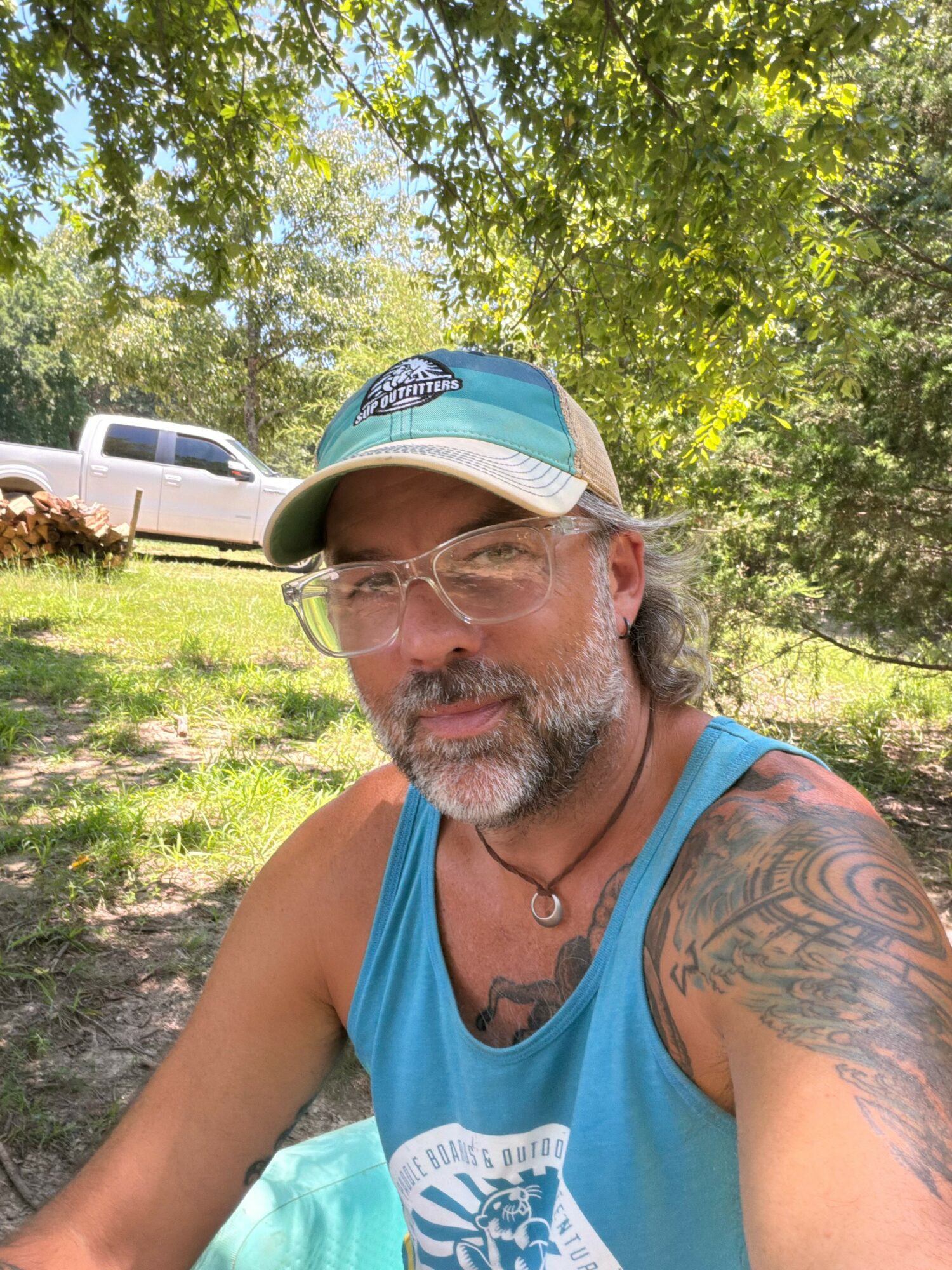 Man with glasses, beard, and tattooed arm outdoors, wearing a blue tank top and cap, with trees and a vehicle in background.