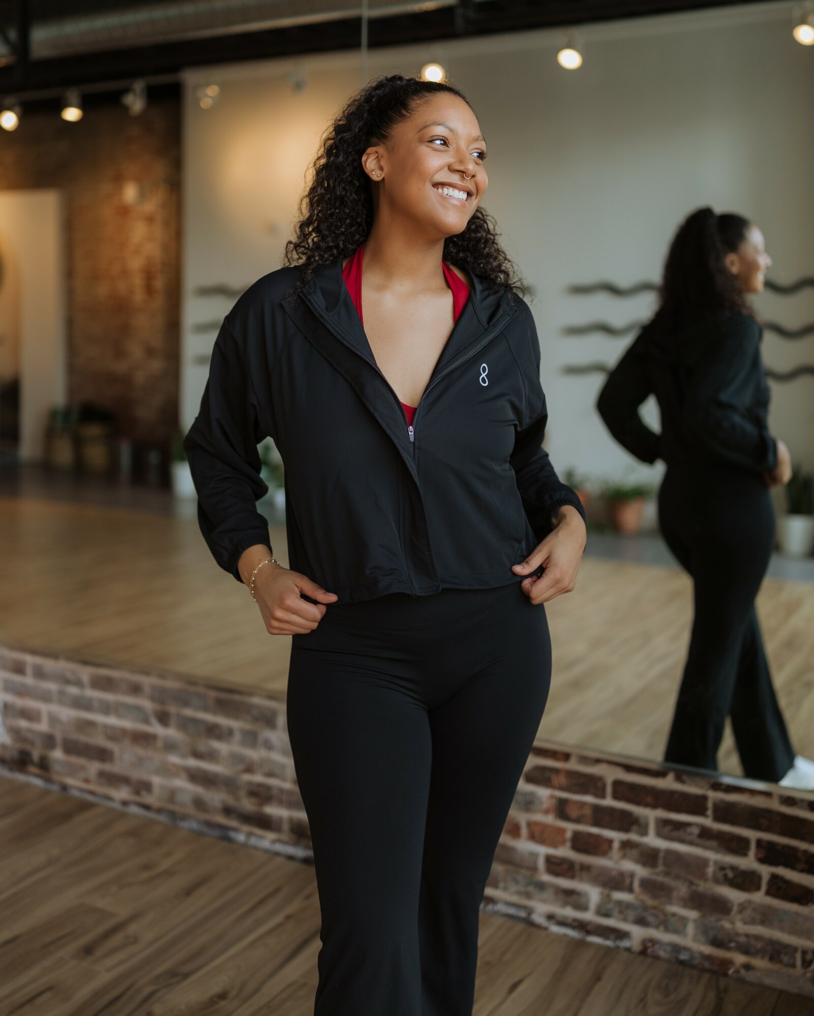 Smiling woman in black athletic wear standing in a gym with a mirror behind her.