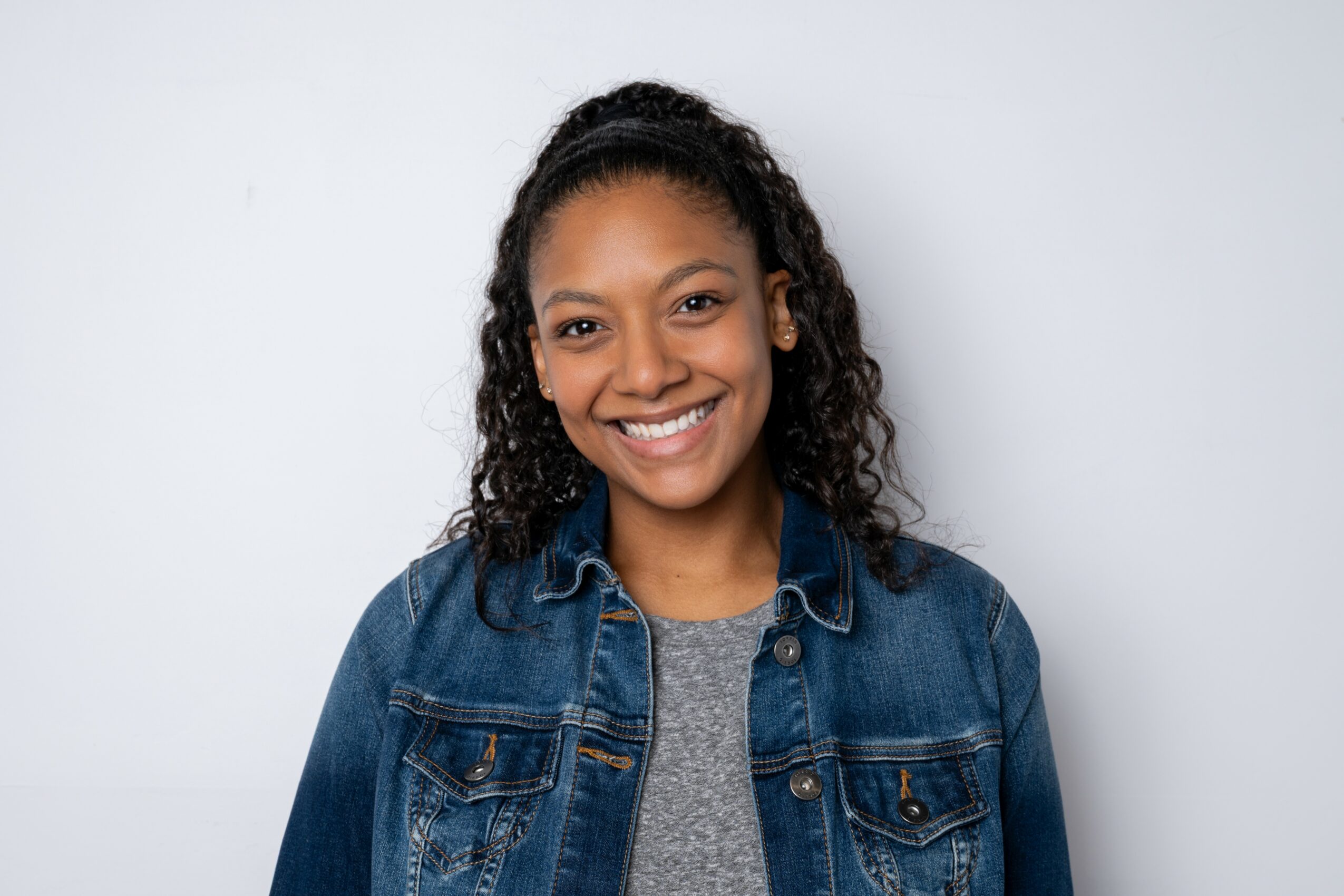 Smiling woman with curly hair wearing a denim jacket and gray shirt against a plain background.