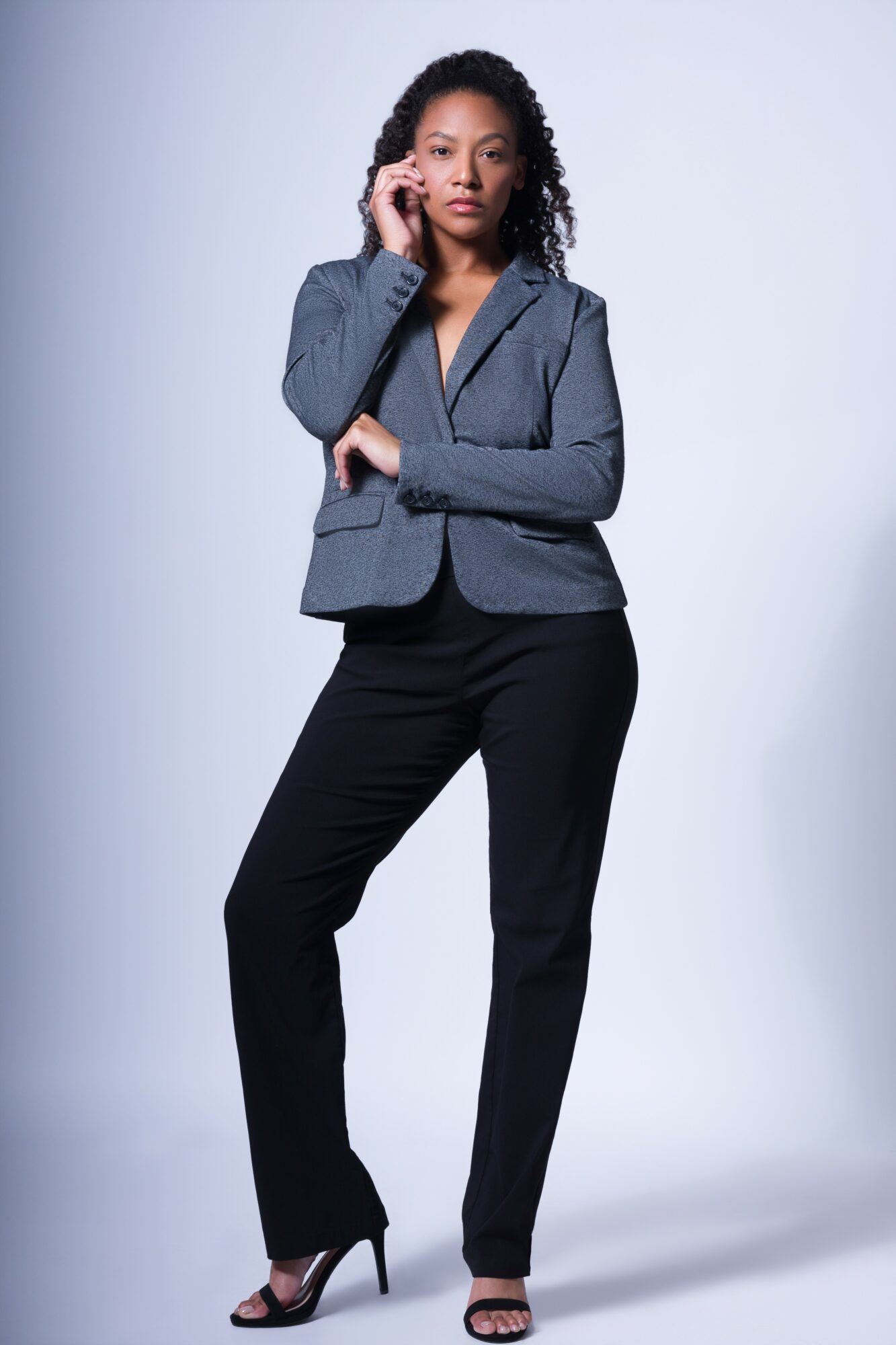 Woman in a gray blazer and black pants standing with one hand on her face, looking at the camera, against a plain background.
