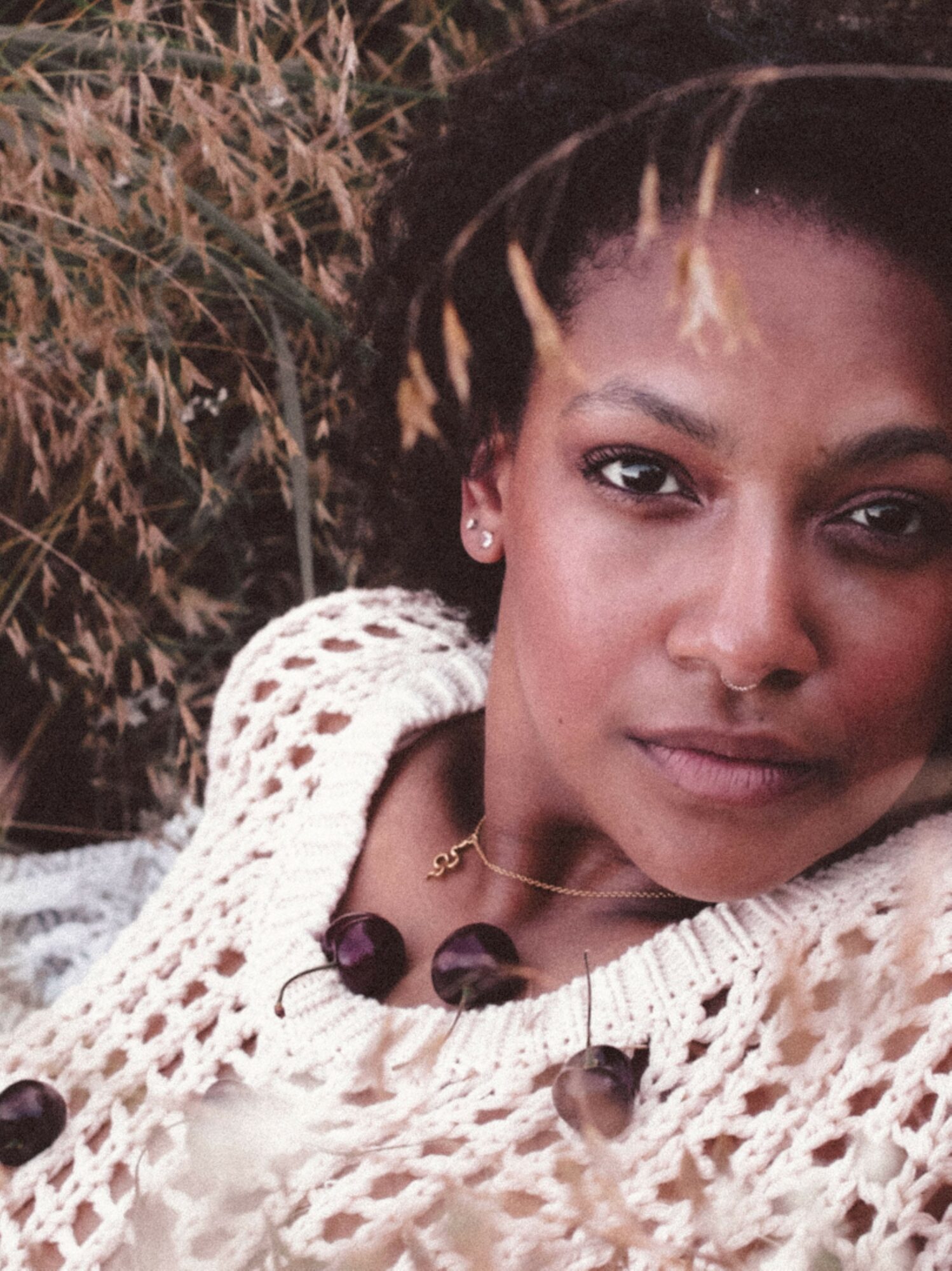 Close-up of a woman with dark hair and earrings, wearing a cream-colored knitted garment, outdoors with dry plants in background.