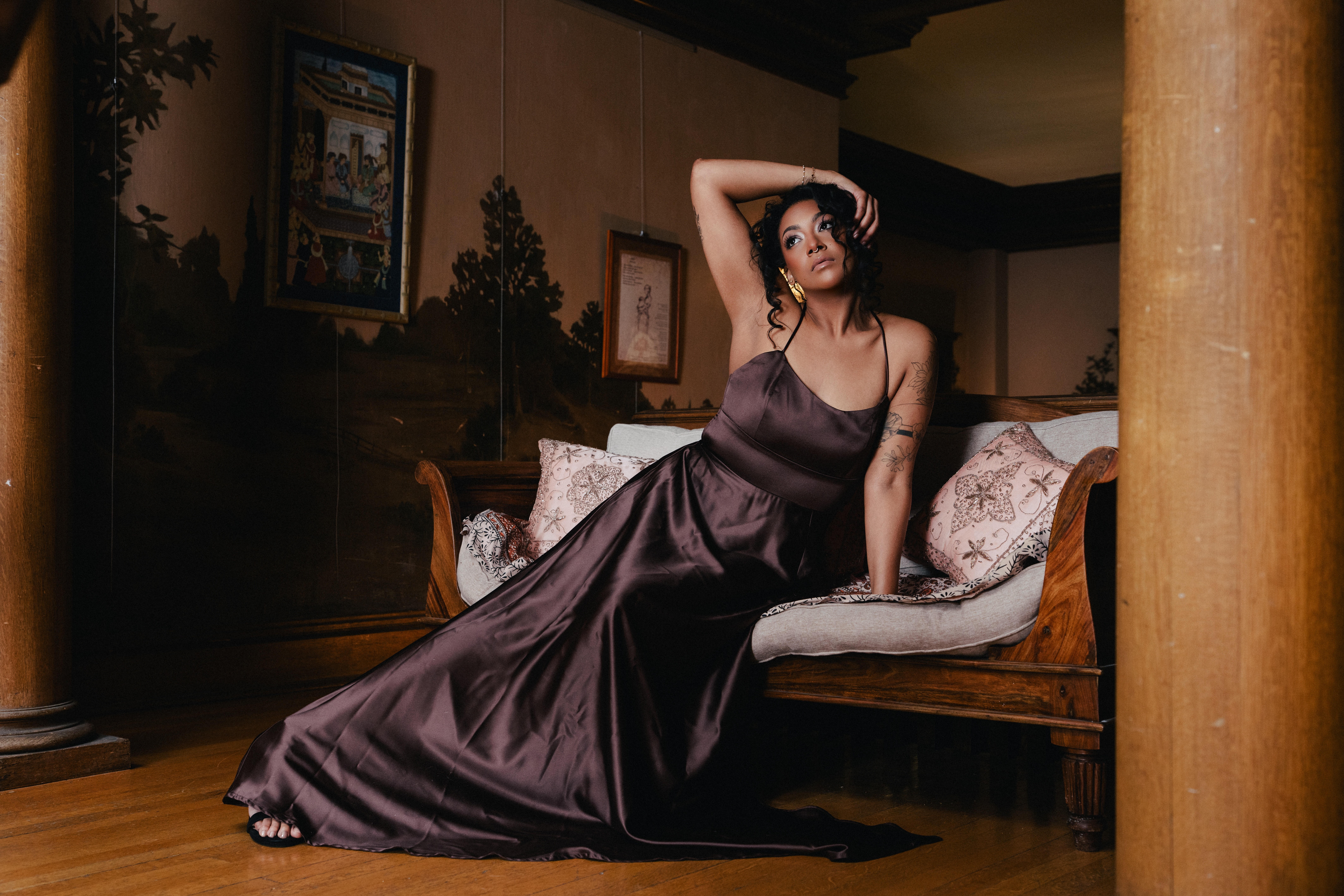Woman in a dark satin gown sitting on a vintage sofa in a dimly lit room with framed pictures on the wall.