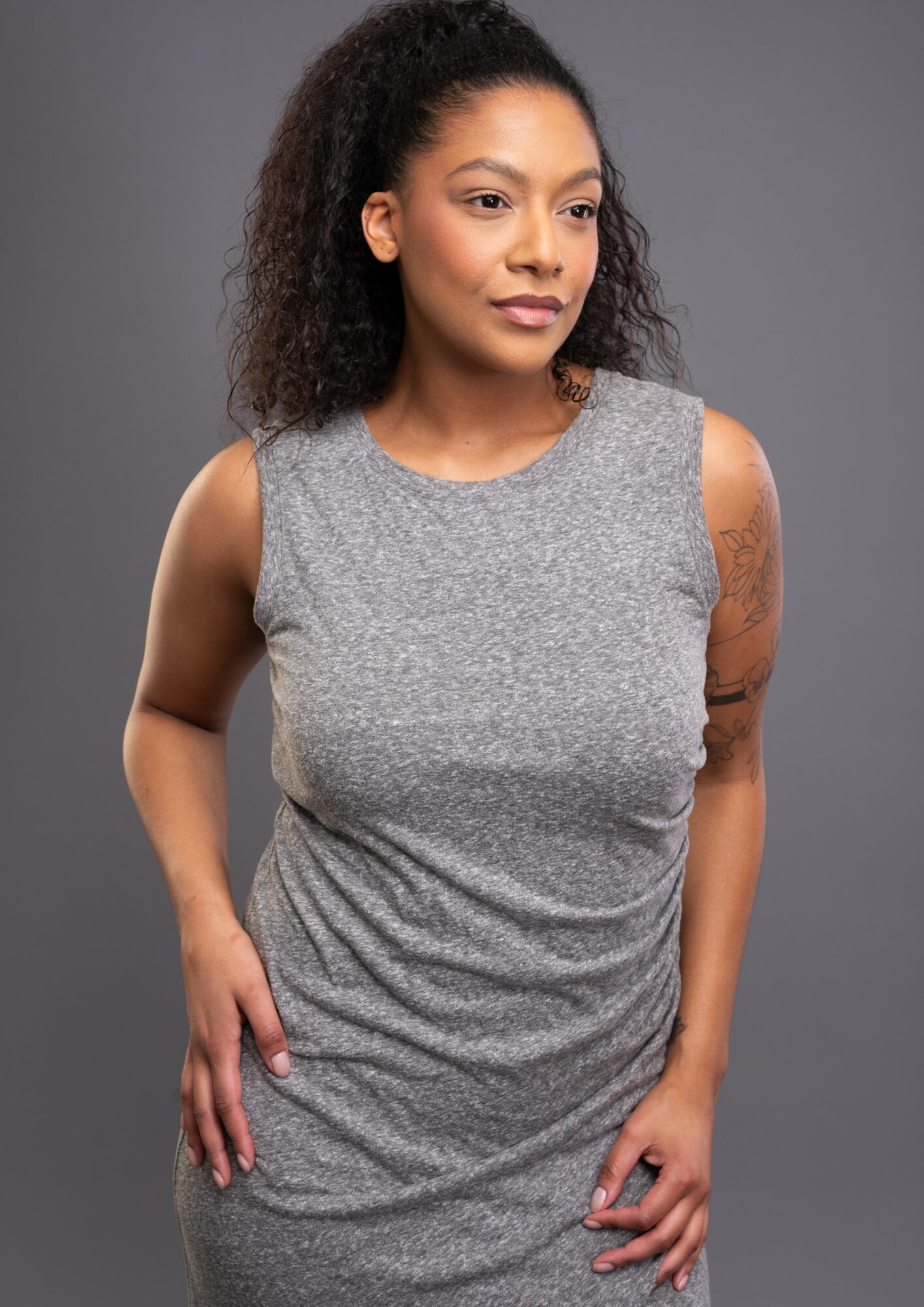 Young woman with curly hair wearing a sleeveless gray dress, standing against a gray background.