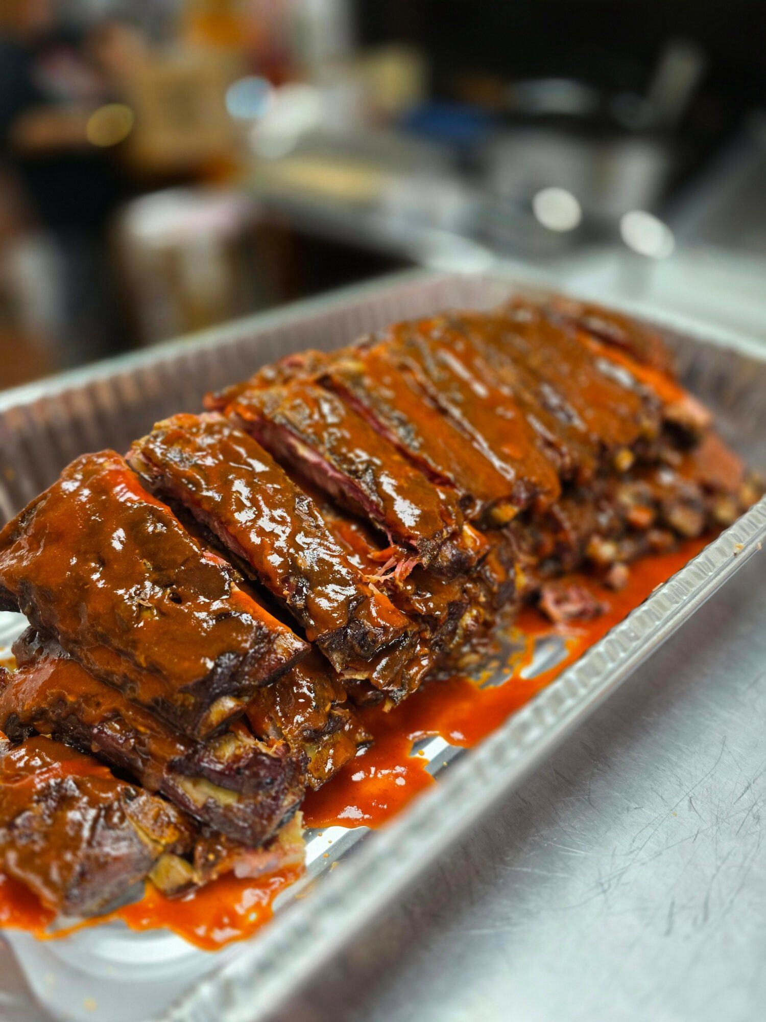 Sliced glazed meatloaf on a metal tray with sauce, in a kitchen setting, with blurred background.