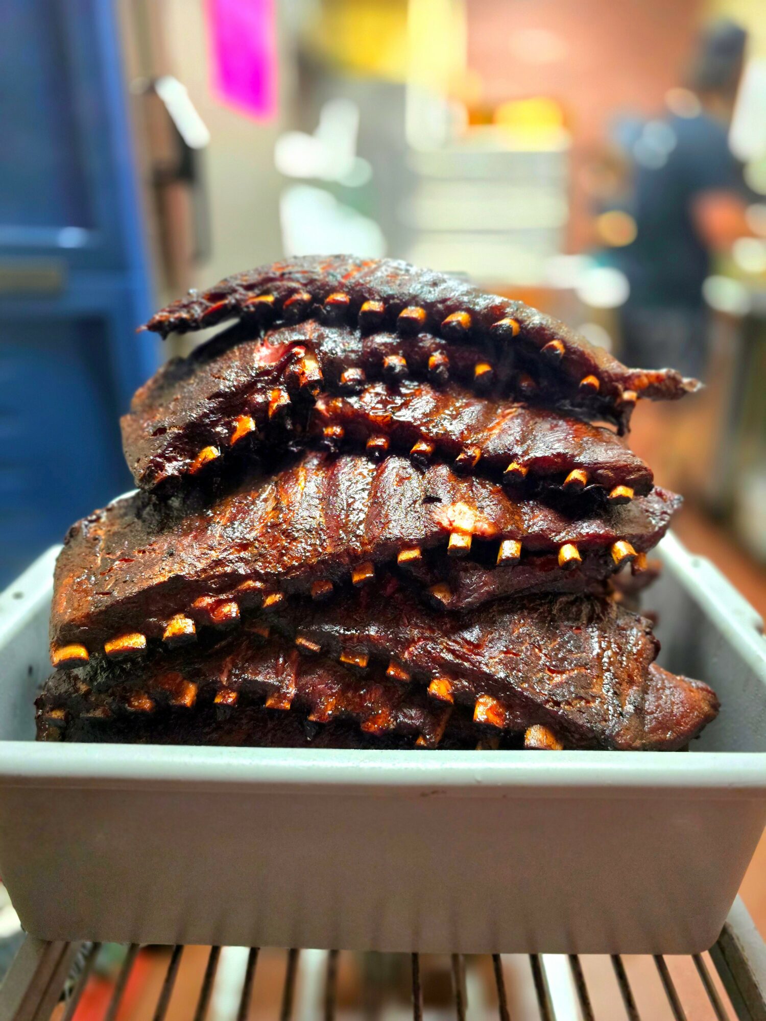 Stack of glazed ribs with visible seasoning and caramelization, in a white container, blurred background of a restaurant or market.